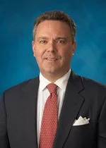 Professional headshot of a man in a suit and tie against a blue background.