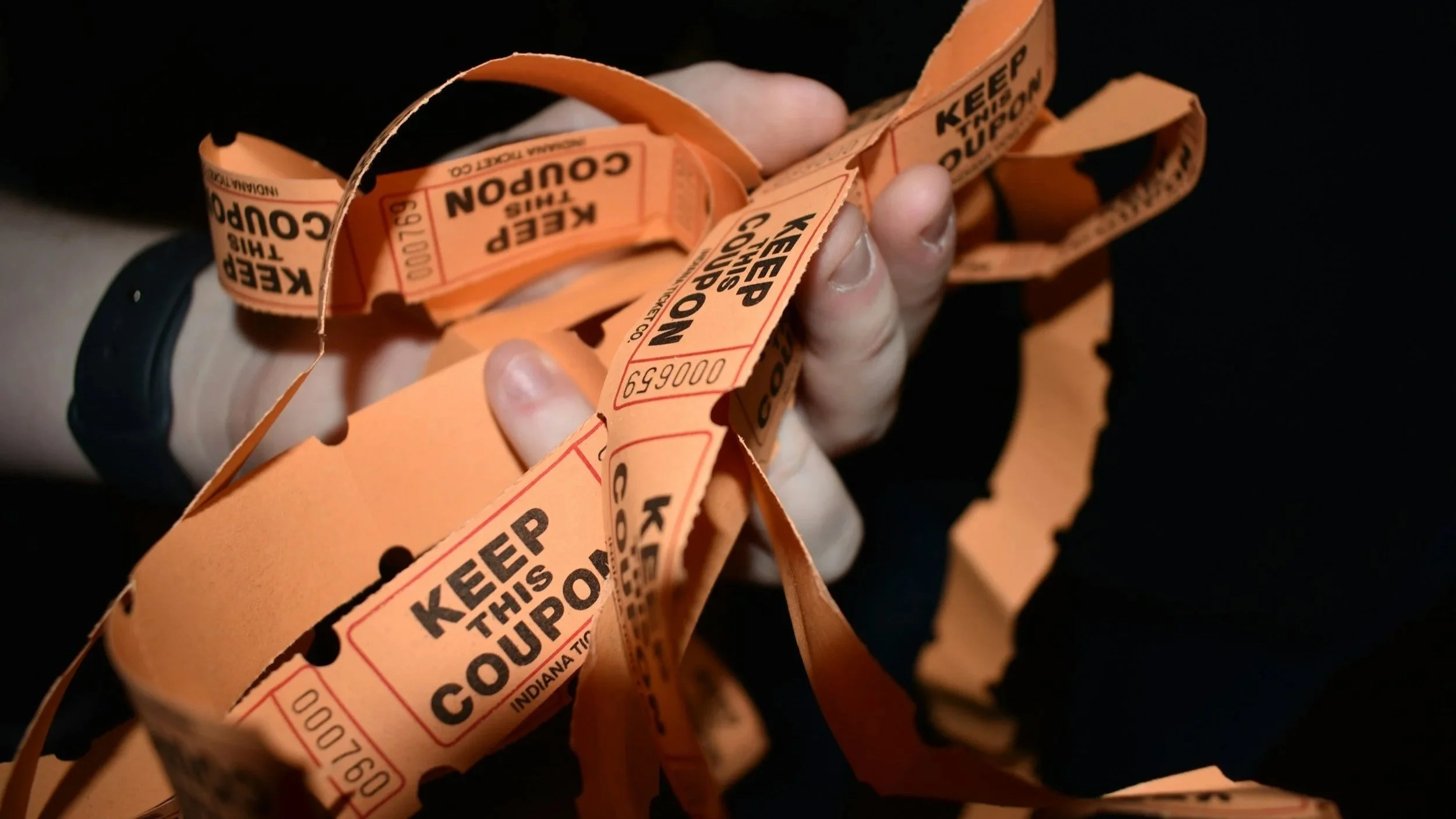 Close-up of a hand holding multiple orange food court tickets with black text that says "Keep this coupon" and "Keen Trip Coupon." The person's hand is visible and they are wearing a black watch, with a dark background.
