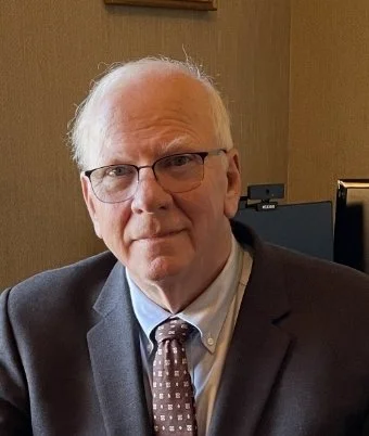 A man with white hair and glasses wearing a suit and tie sitting indoors.