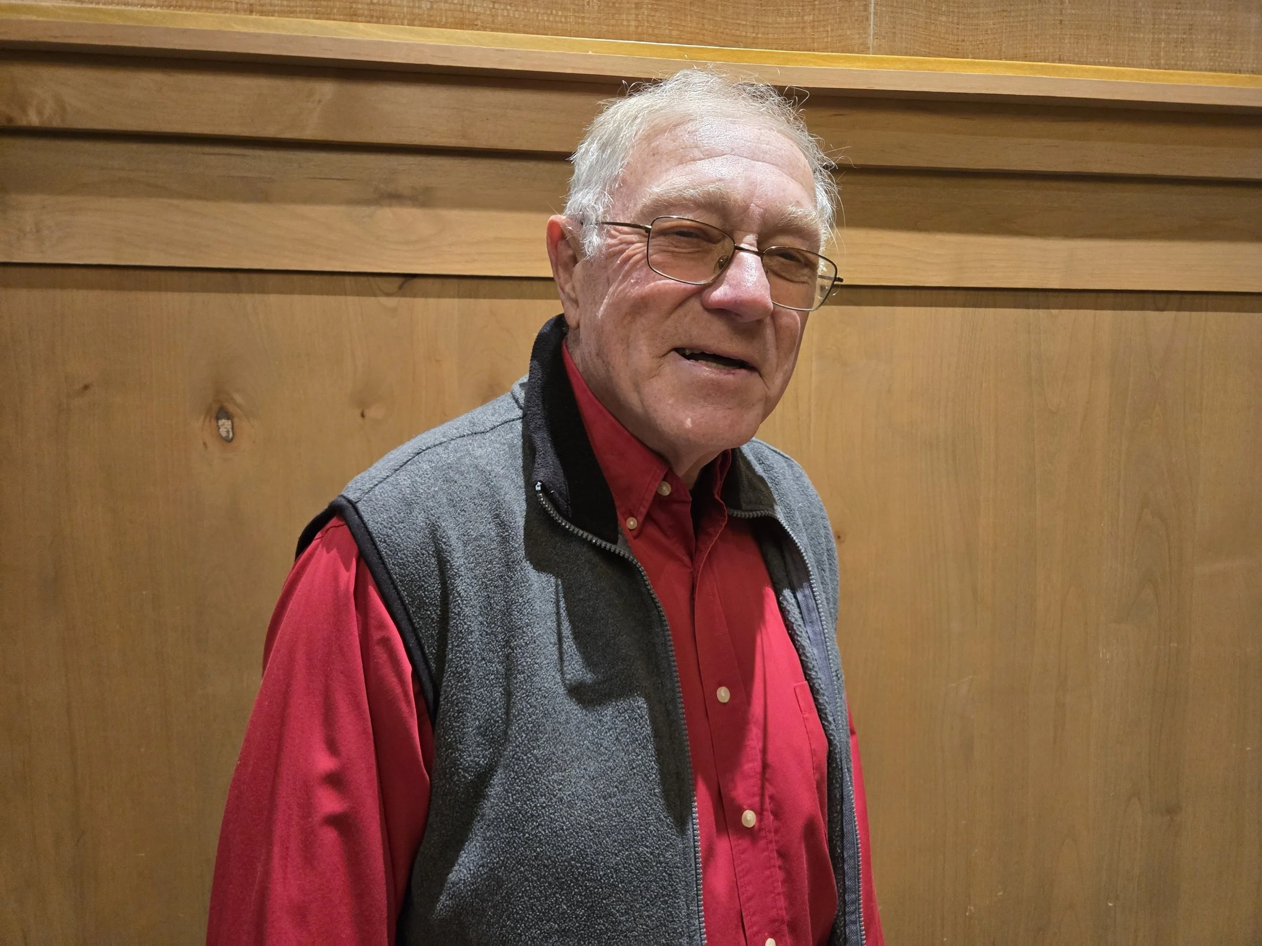 Older man with glasses and gray hair smiling, wearing a red shirt and a gray vest, standing in front of a wooden wall.