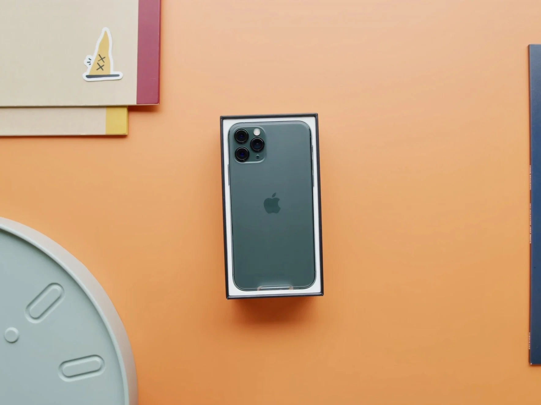 An iPhone in its box on a peach-colored surface, surrounded by books and other objects.