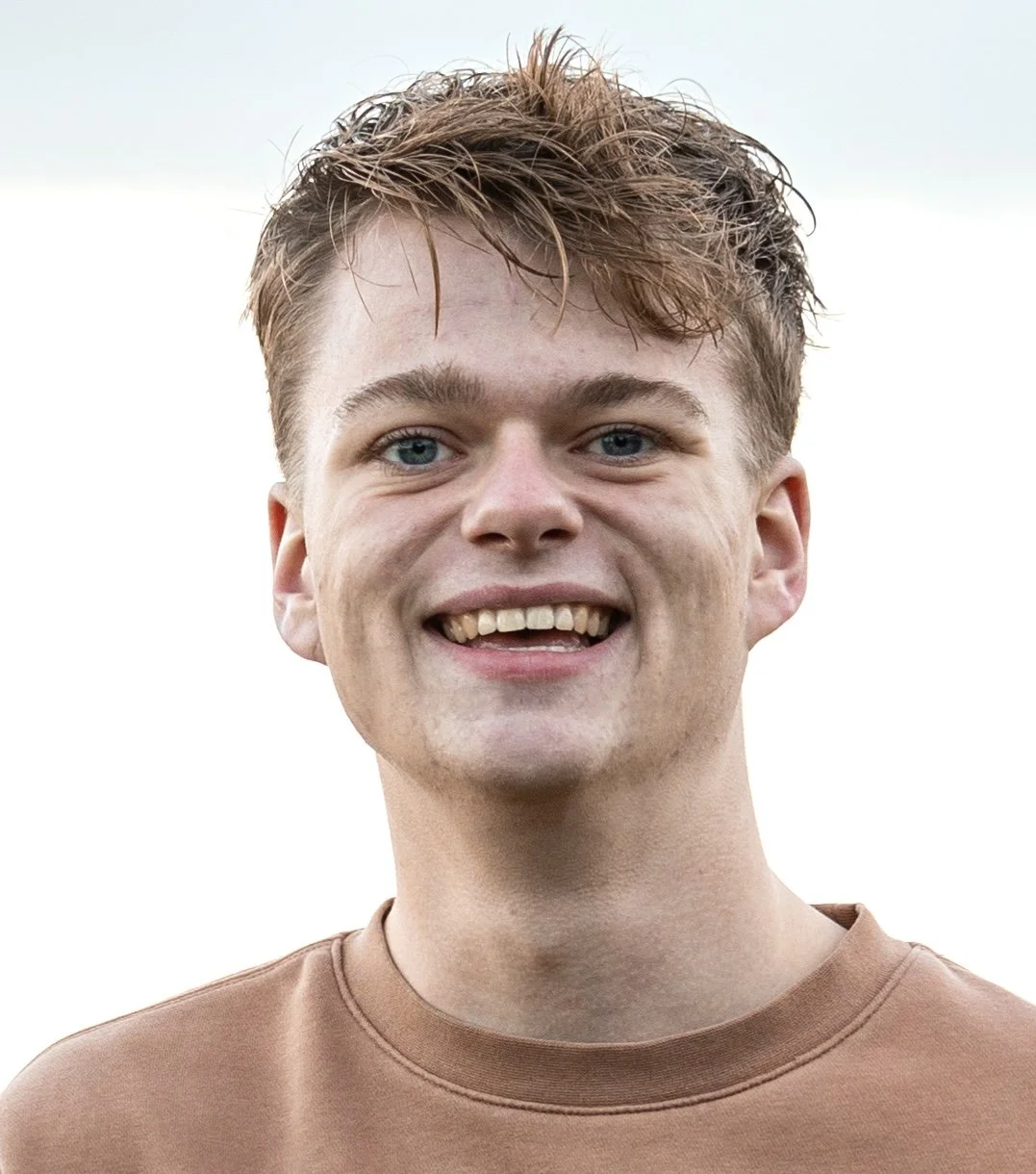 A smiling young man with short light brown hair, blue eyes, and fair skin, wearing a brown shirt, outdoors with a cloudy sky background.