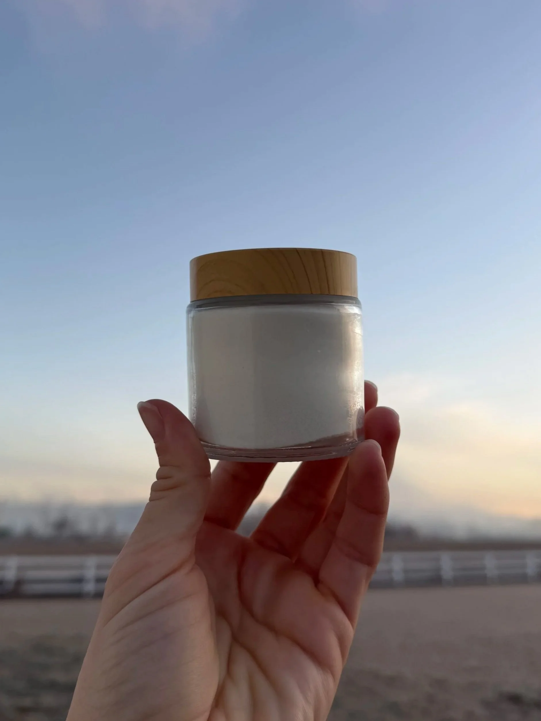 A hand holding a clear glass jar with a wooden lid, containing white balm, against a backdrop of a sky at either dawn or dusk, with a fence and trees in the distance.
