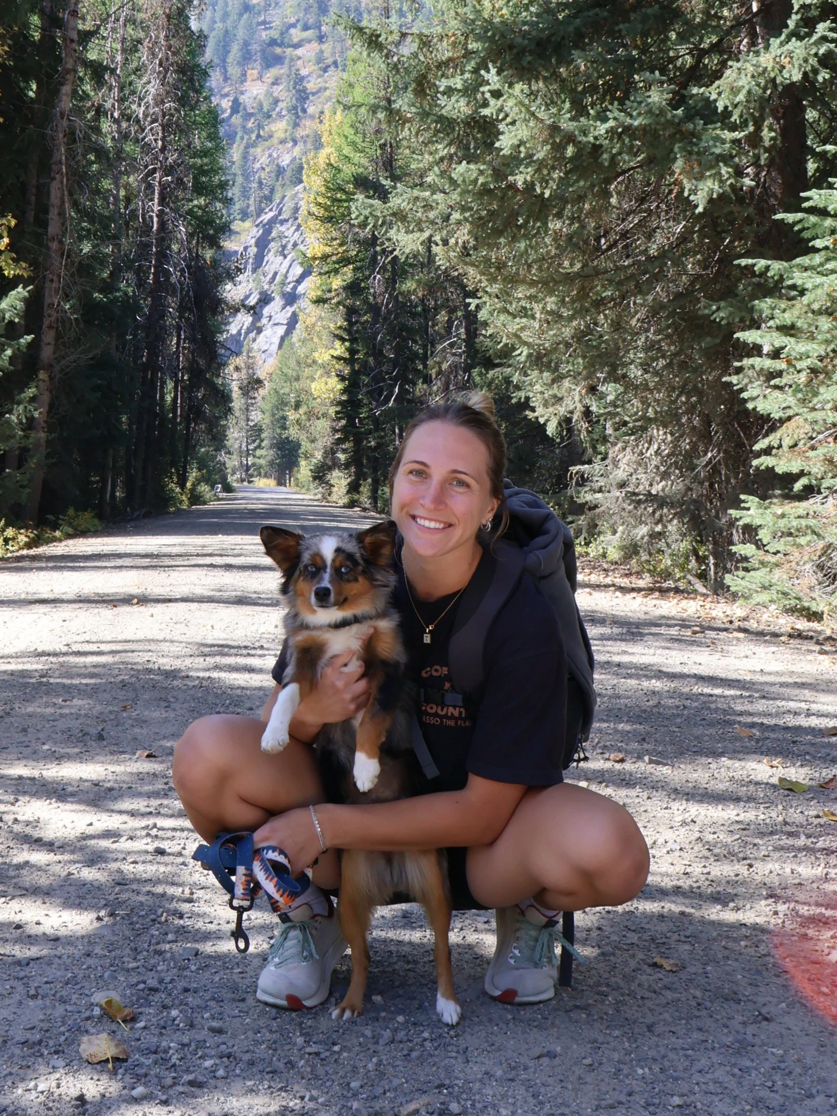 A woman smiling and kneeling on a dirt trail in a forest, holding her small dog, surrounded by tall trees with a mountain in the background.