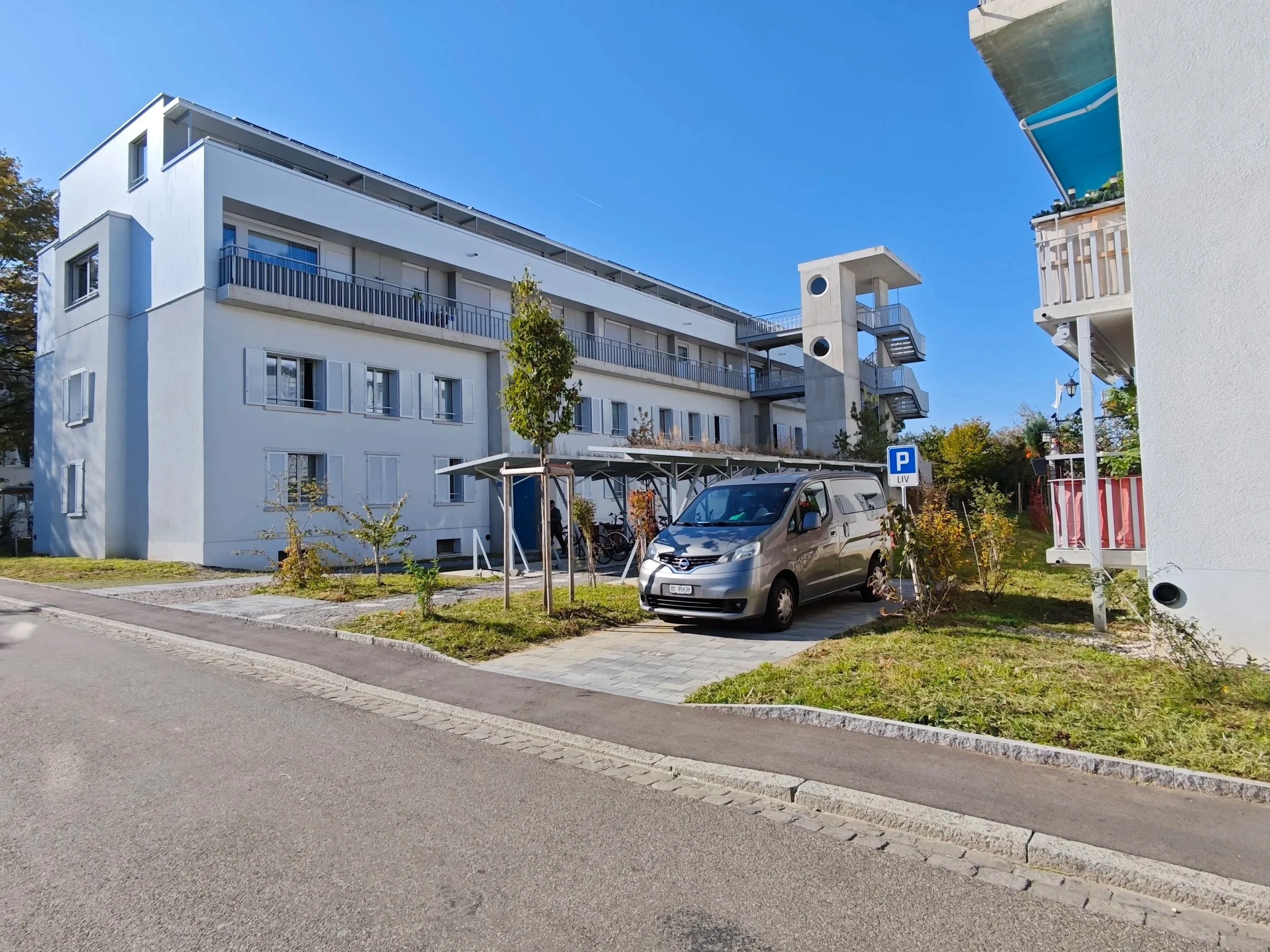 Mehrstöckiges weißes Wohngebäude mit Balkon, Parkplatzzone mit Auto, Fahrradständer und kleinen Bäumen vor dem Haus.