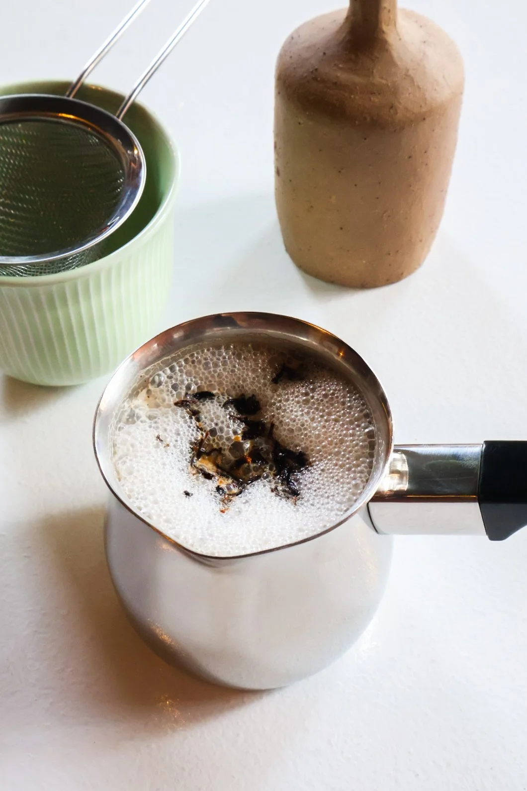 A pour-over kettle of brewed chai, a green cup with a metal filter, and a brown bottle on a white surface.