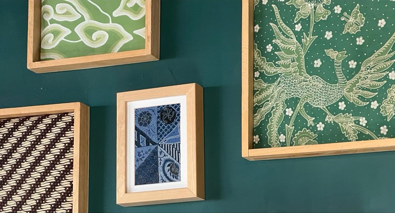 Gallery wall with four framed batik art pieces in wooden frames, including a green and white abstract pattern, a detailed green and white floral and bird pattern, a black and white striped pattern, and a dark blue pattern with geometric designs.