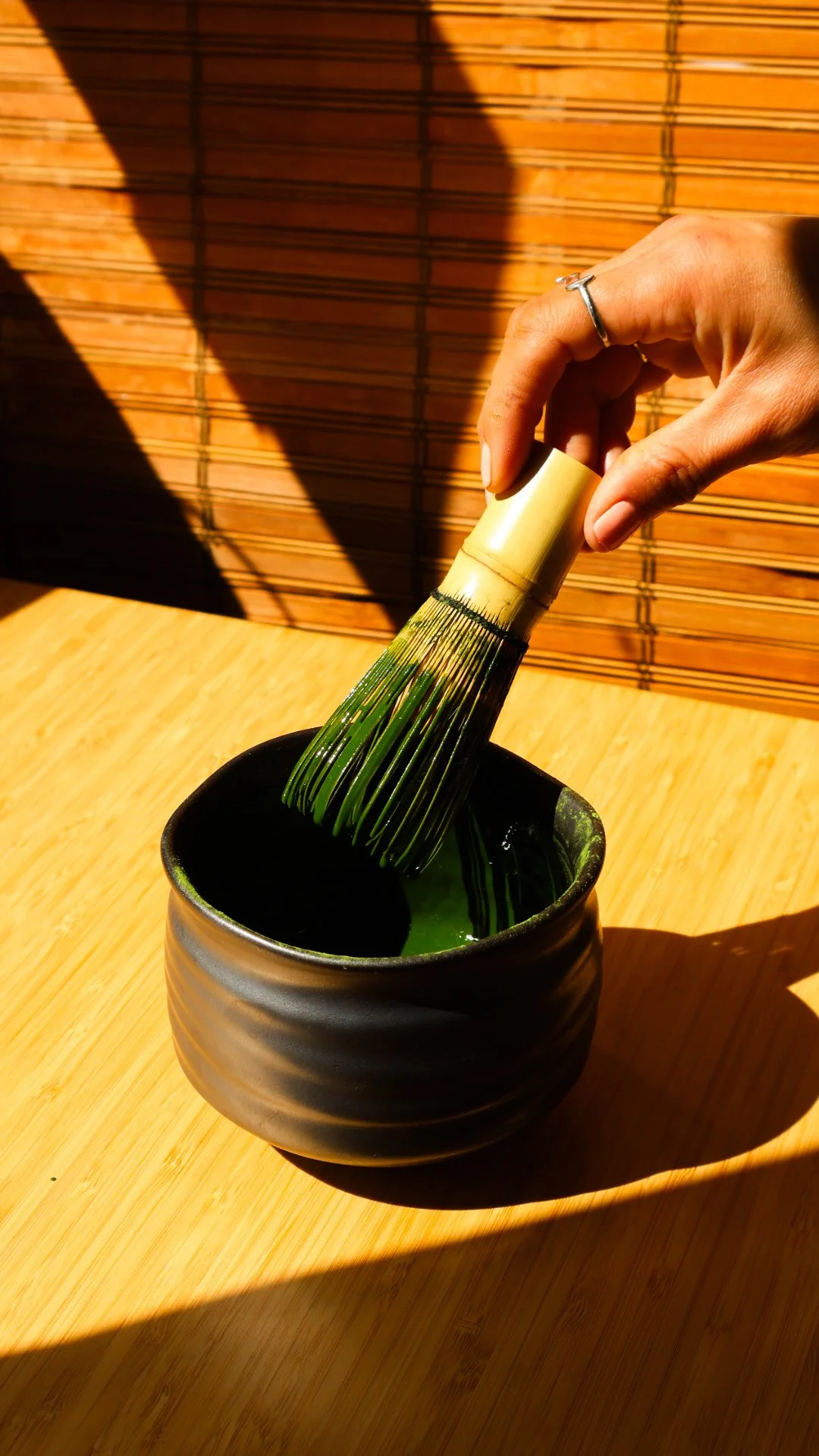 A hand holding a matcha whisk over a black cup with freshly whisked matcha inside