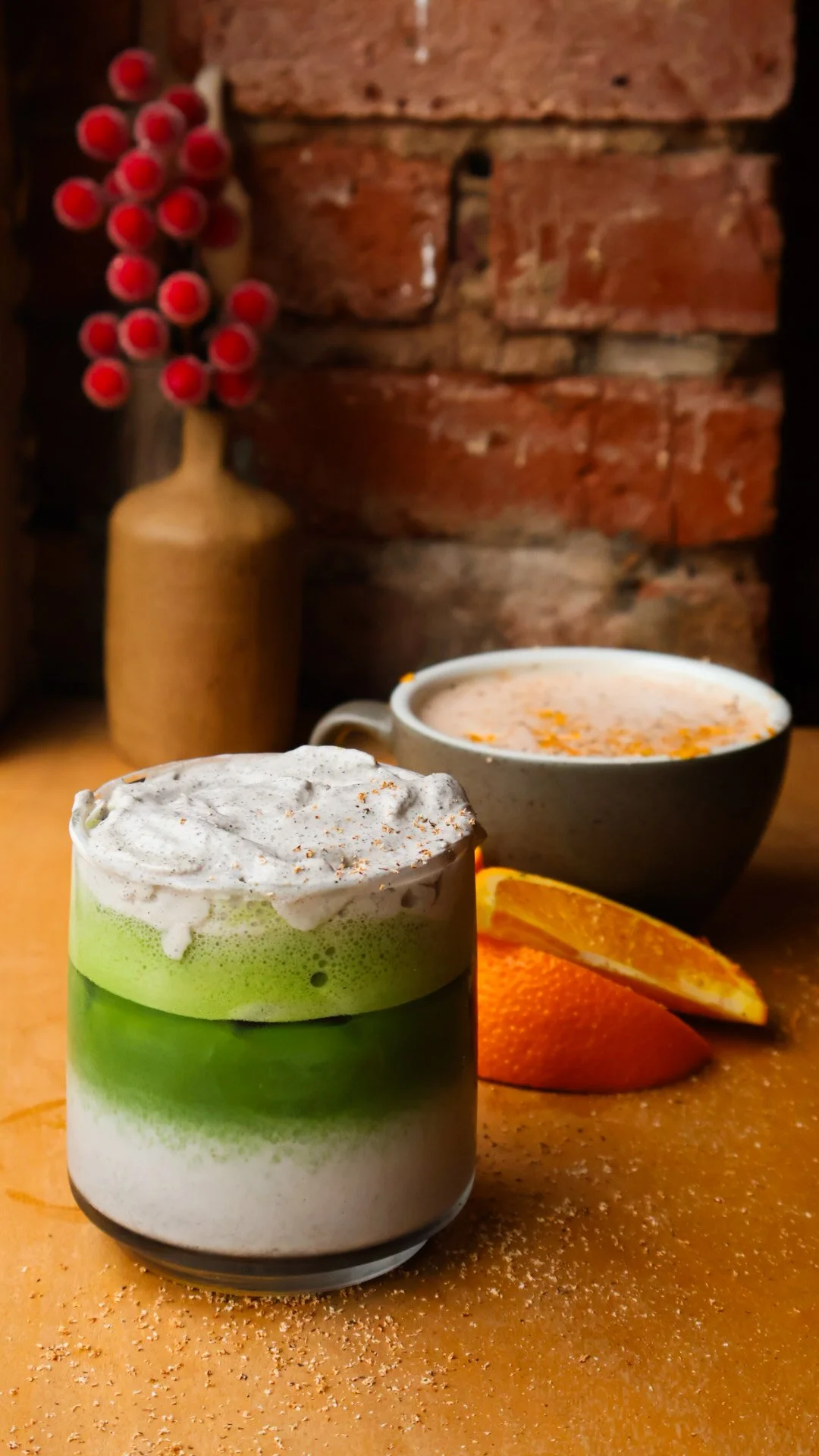 A green and white iced matcha drink an a mug of hot chocolate on a brown countertop with orange slices between them.