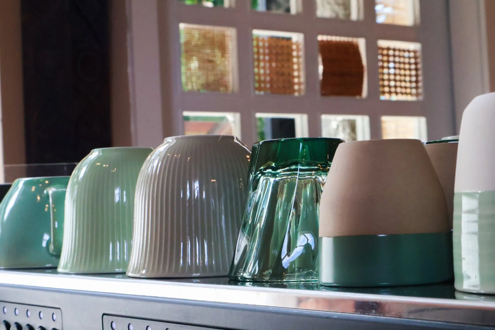 Various ceramic cups and bowls in white, green, and cream colors arranged on a kitchen counter with a window in the background.