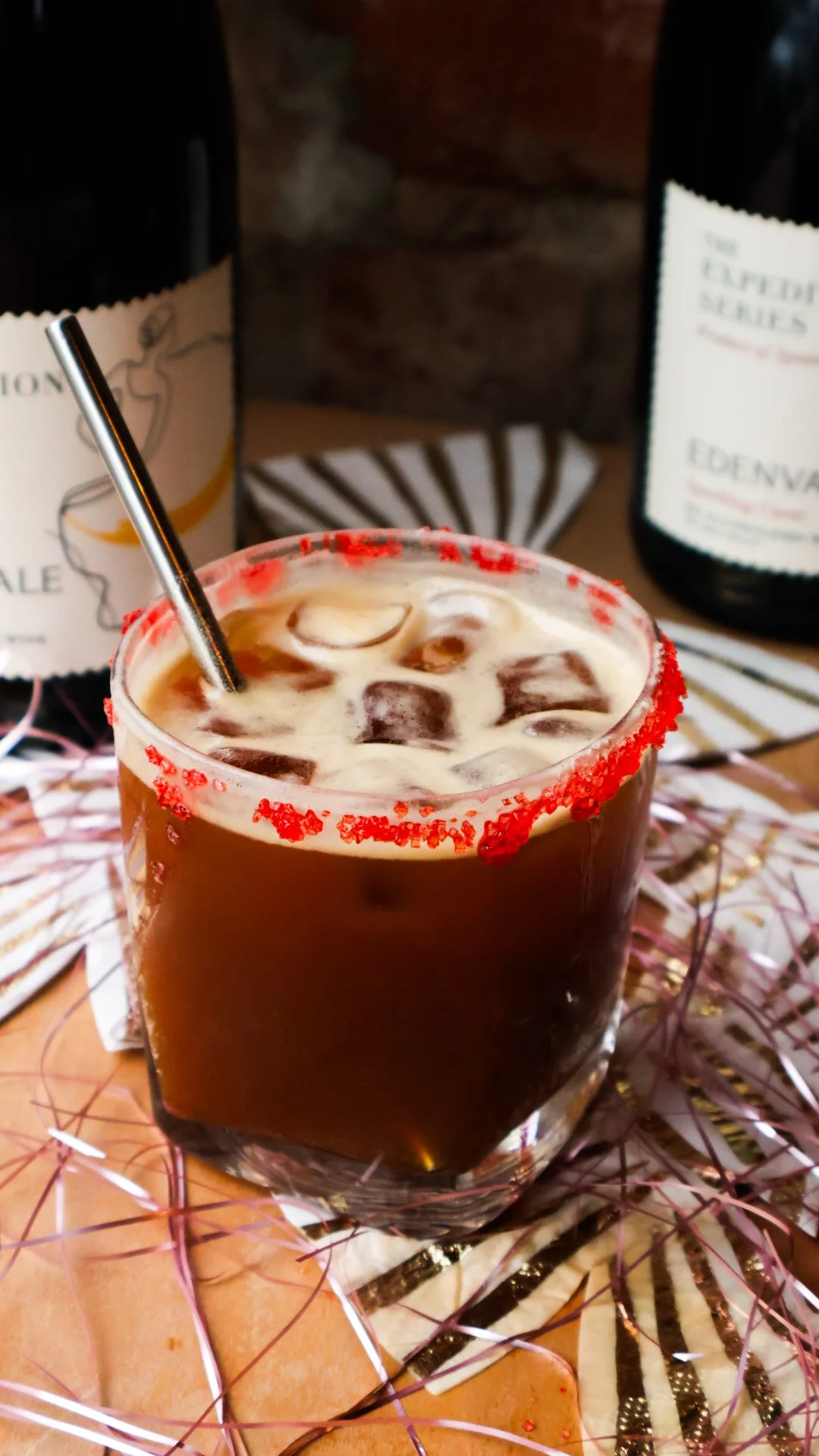 A iced drink with red sugar crystals around the rim and a steel straw. Two bottles of a non-alcoholic beverage rest behind.