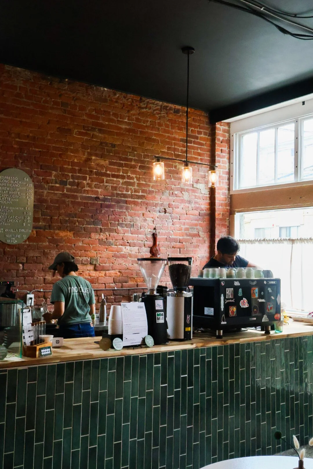 Interior of a coffee shop with a brick wall, a counter with coffee-making equipment, a menu board, and the Wairua owners working. Large window letting in natural light.