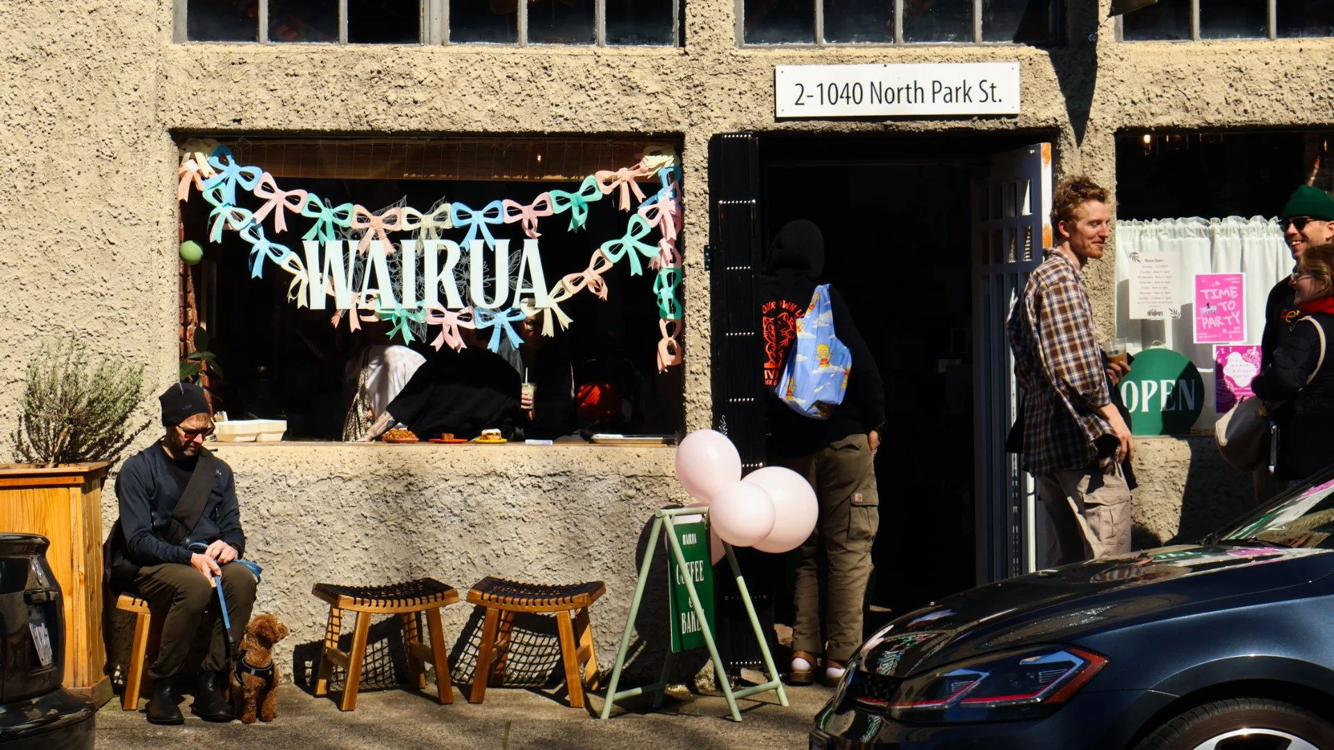 The front entrance to Wairua. Colorful streamers in the window. A customer sits outside with their dog, and others chat nearby.