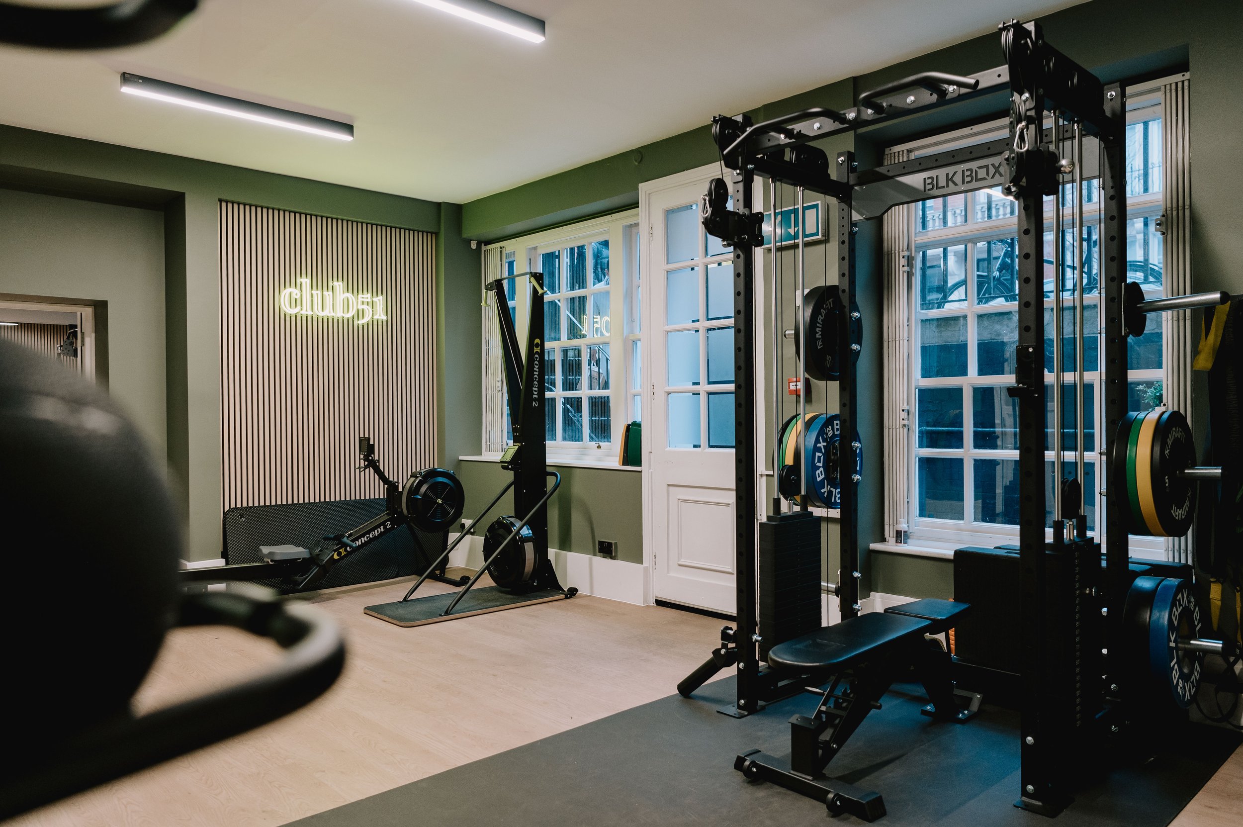 Modern indoor gym with weight lifting equipment, rowing machine, and a neon sign that says 'club 51' on the green wall.