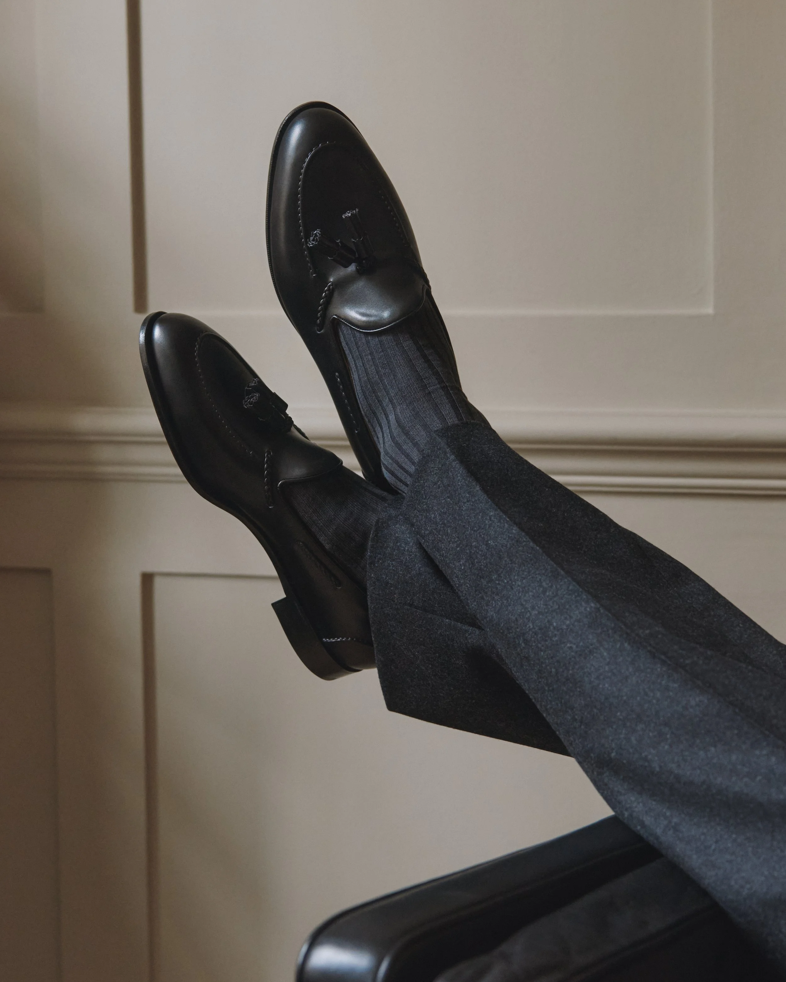 Person wearing black dress shoes, dark socks with stripes, and gray dress pants, with legs extended and resting on a chair in front of a beige wall.