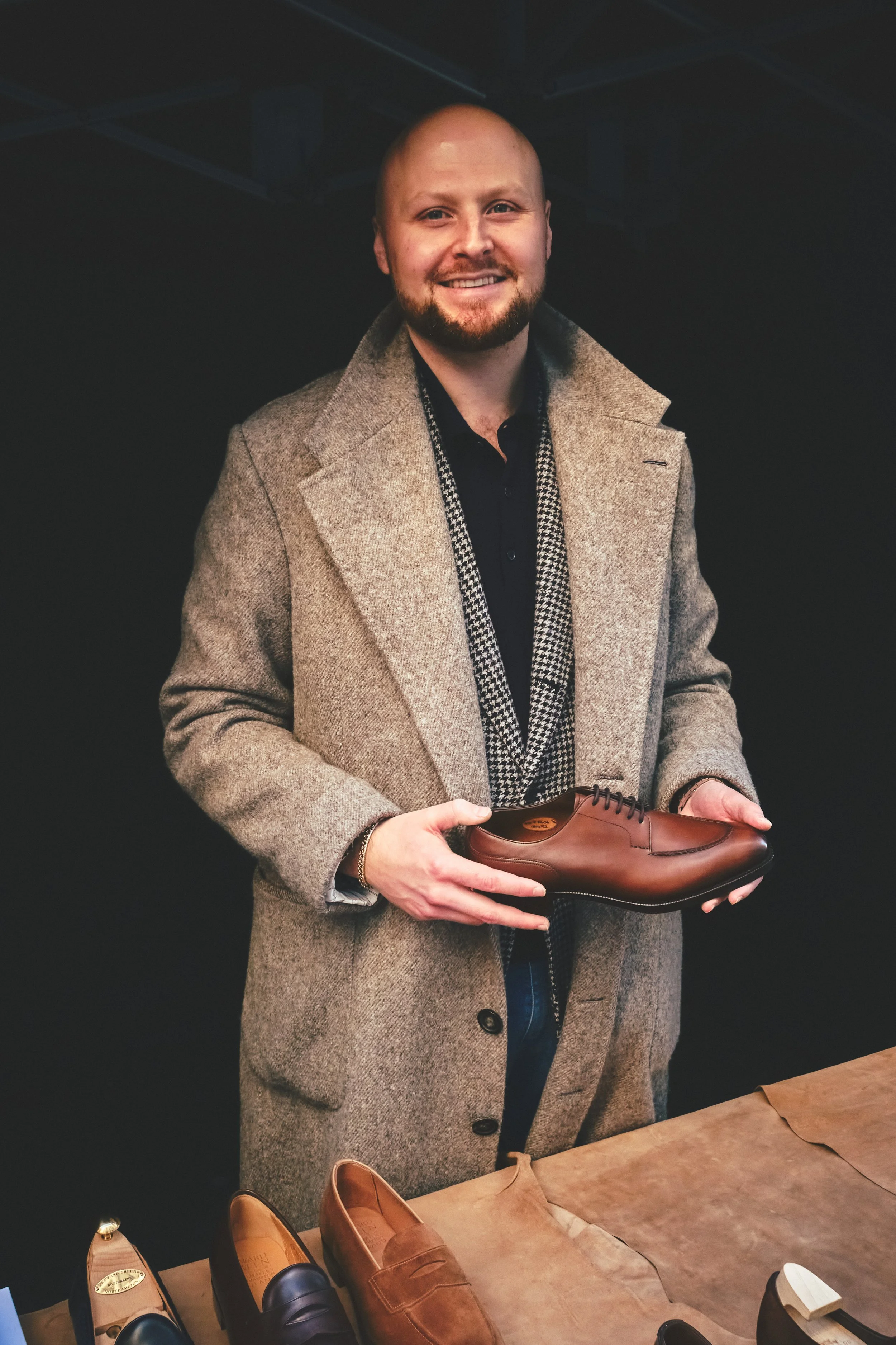 A man in a beige coat holding a brown leather dress shoe, standing behind a table with various shoes.