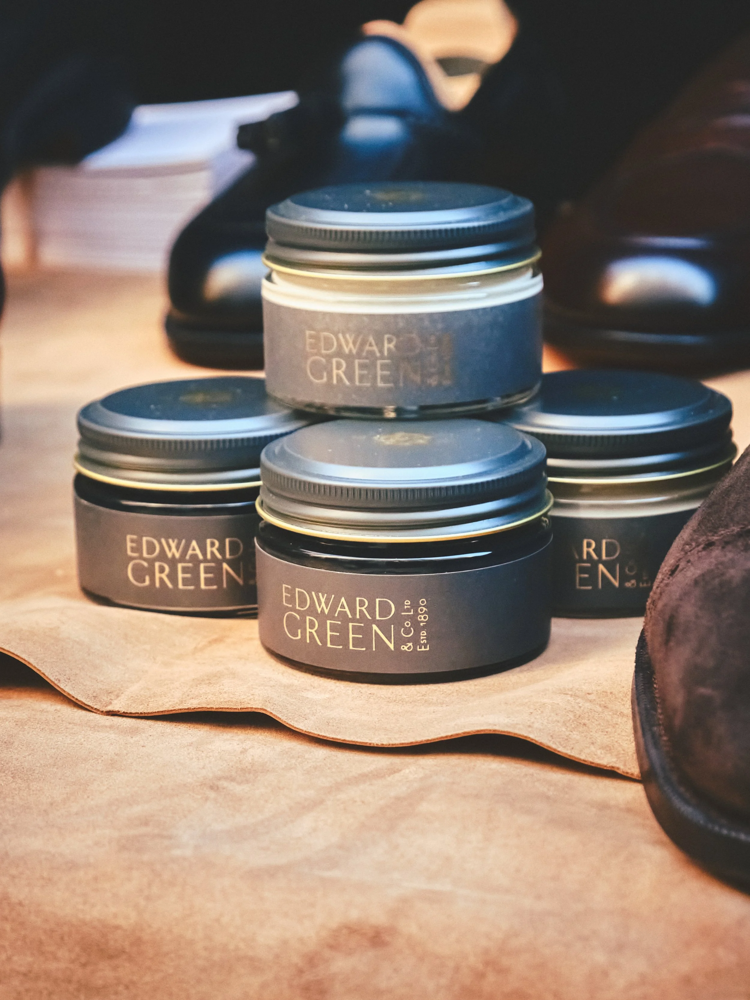 Stack of black jars with black lids labeled 'Edward Green' on a wooden surface, with men's shoes and papers in the background.