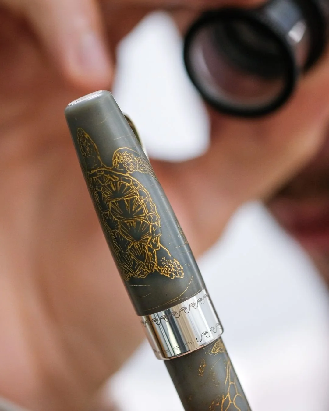 Close-up of a person's face with a sneaker and a pen held near their nose. The pen has a decorative design with a butterfly and intricate gold detailing.