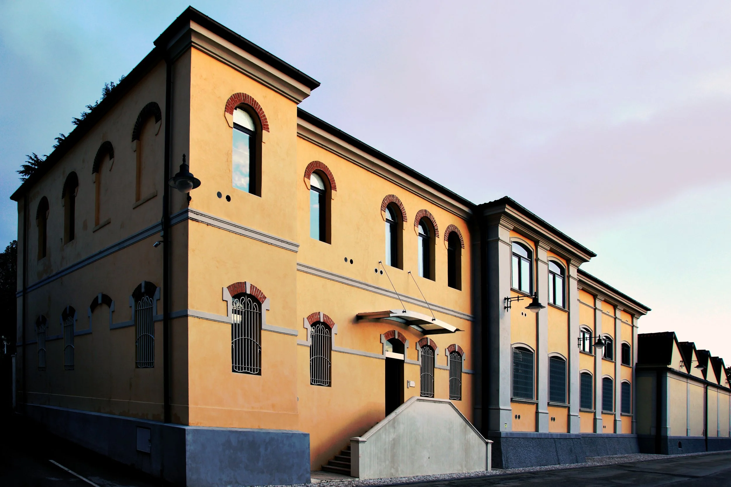 A multi-story yellow building with arched windows and decorative brick accents, situated on a street at dusk.