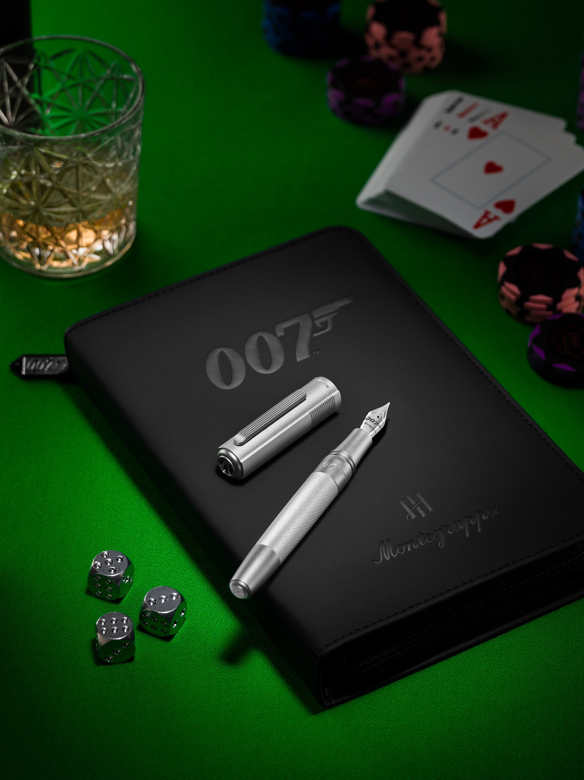 A black Montegrappa notebook with a 007 James Bond logo on it, a silver pen, a lighter, three silver dice, a glass of whiskey, playing cards, and poker chips on a green poker table.