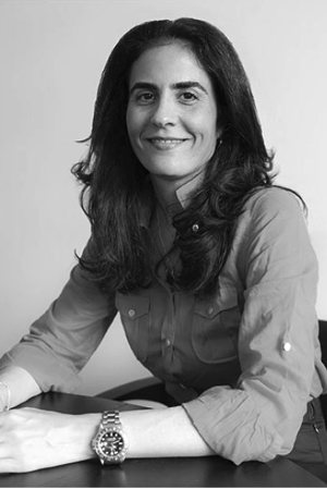 Black and white photo of a woman with long, wavy hair smiling, sitting at a table with her arms resting on it.
