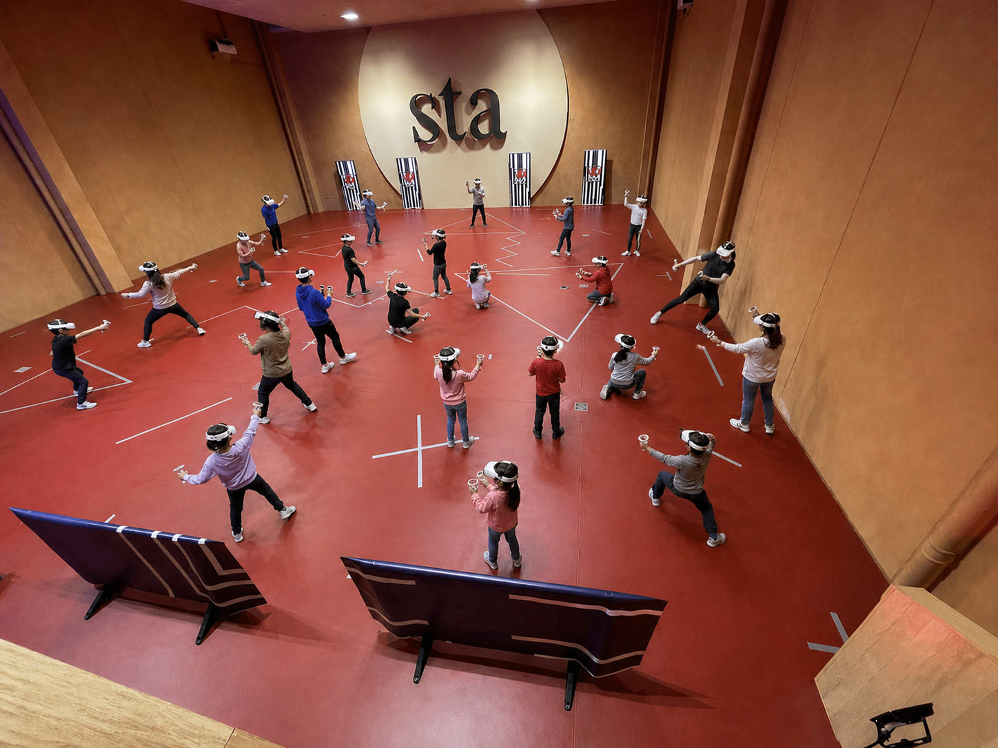 Muchos niños en una cancha con pantallas y un muro grande con la palabra 'sta' en el fondo, participando en una actividad con gafas de realidad virtual.