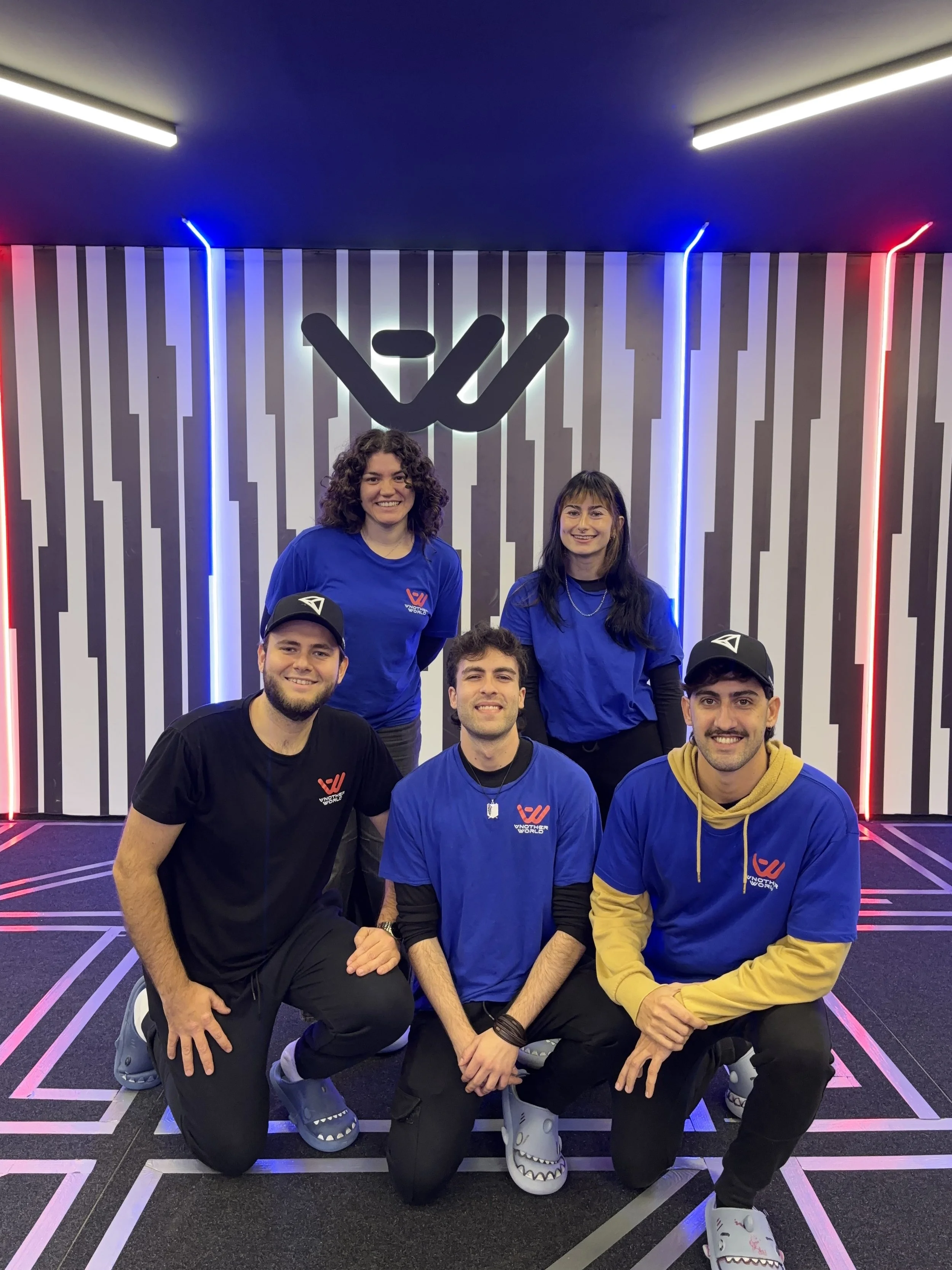 Grupo de seis personas sonriendo y posando en un espacio con decoración moderna y luces de neón, algunos llevan camisetas con un logo que dice 'WONTHY WORLD'