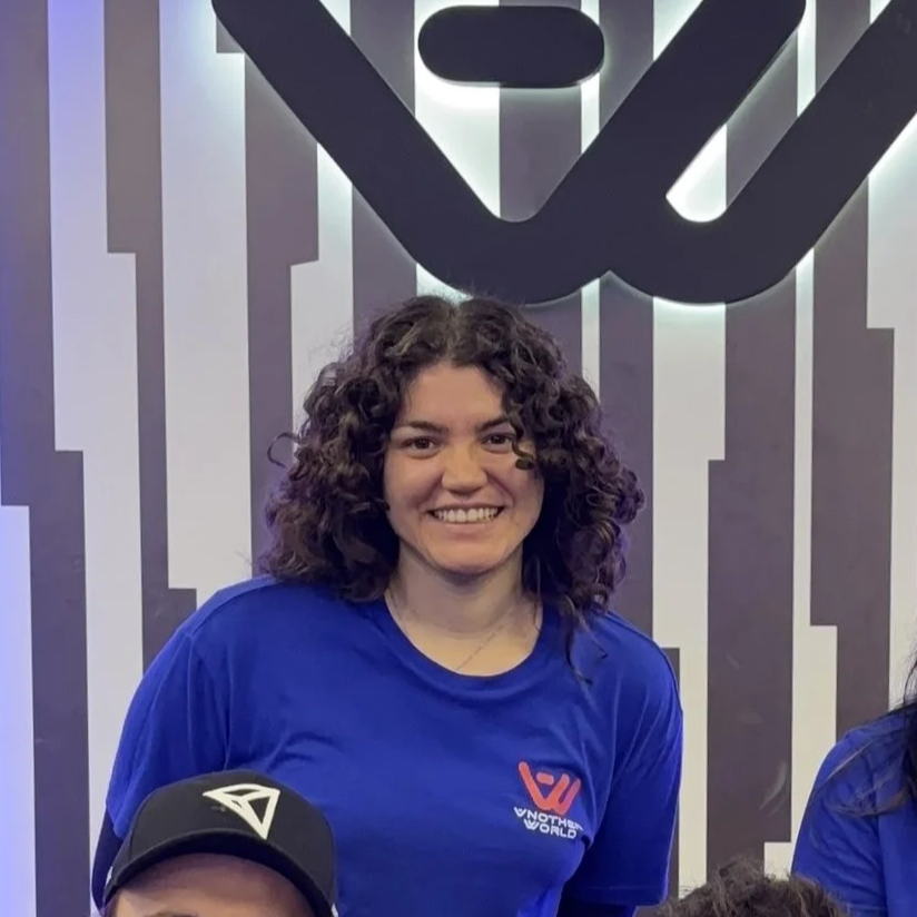Una mujer joven con cabello rizado y sonrisa, vestida con camiseta azul, en un lugar con un fondo de paredes con rayas blancas y negras y un logo grande en la pared.