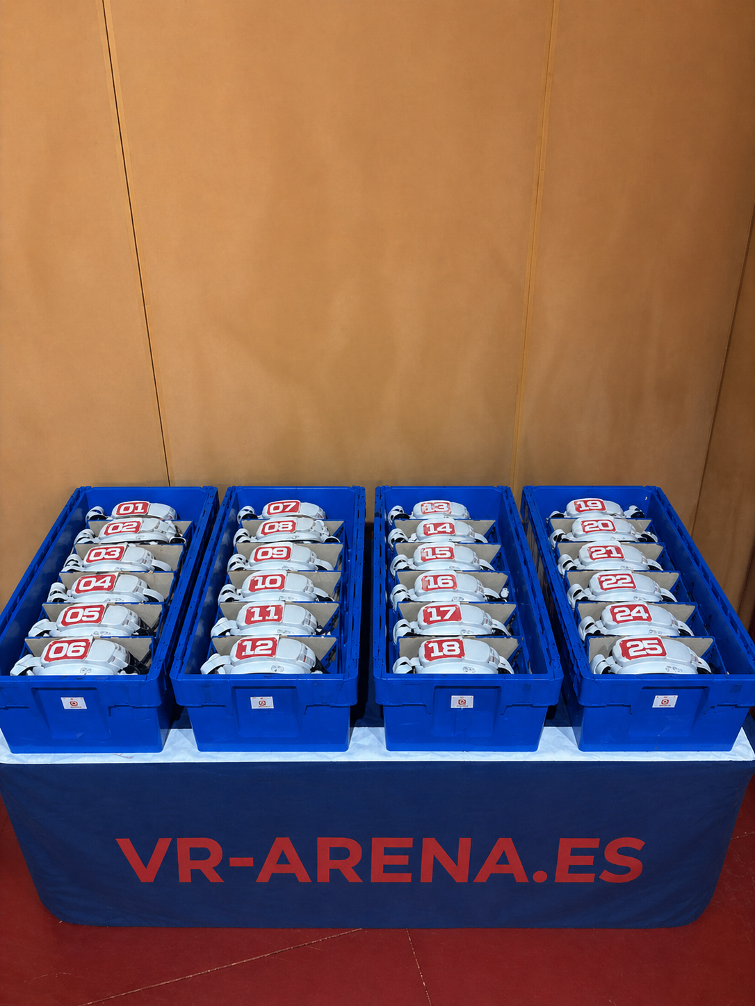 Casco de realidad virtual (VR) en cuatro cajas azules, numerados del 01 al 25, sobre una mesa cubierta con una tela azul que tiene el texto 'VR-ARENA.ES'.