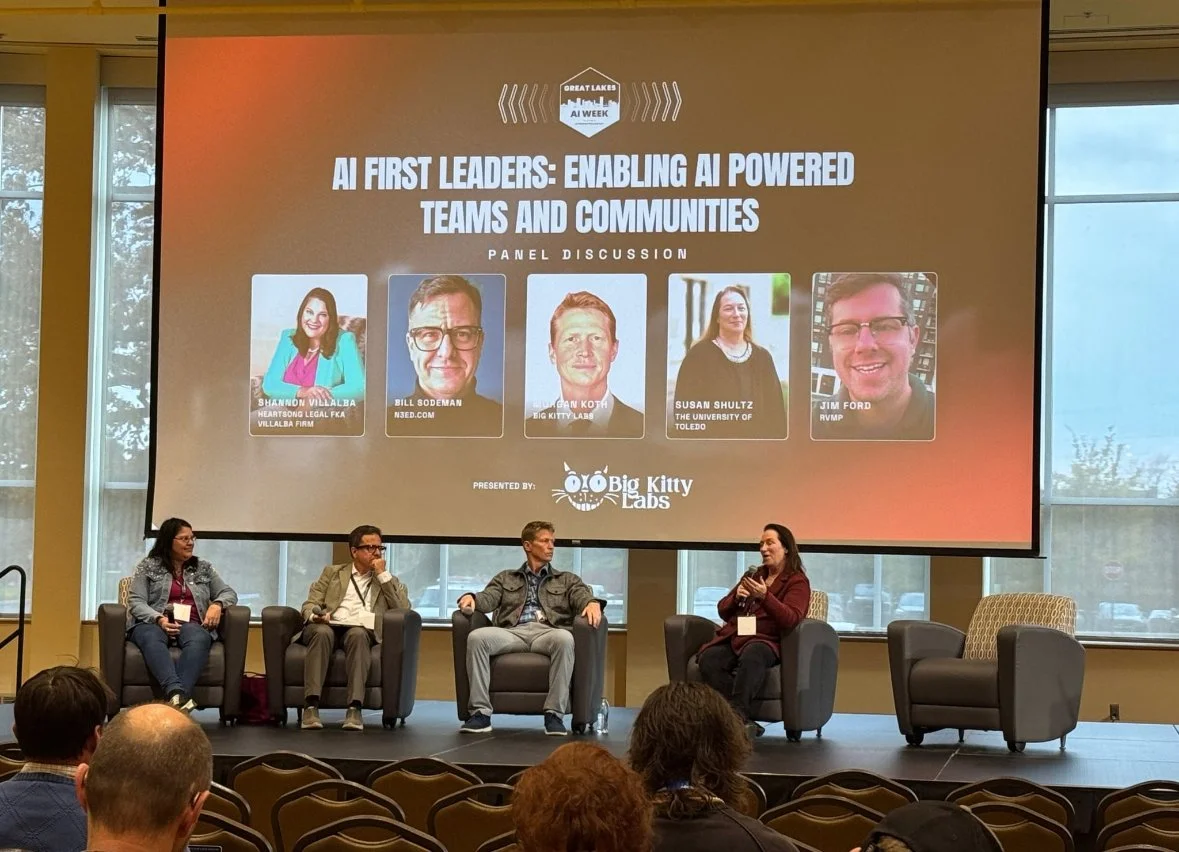 Panel discussion at Great Lakes AI Week. Five panelists sit on stage in armchairs; large screen behind them displays event title and panelist photos with names and affiliations.