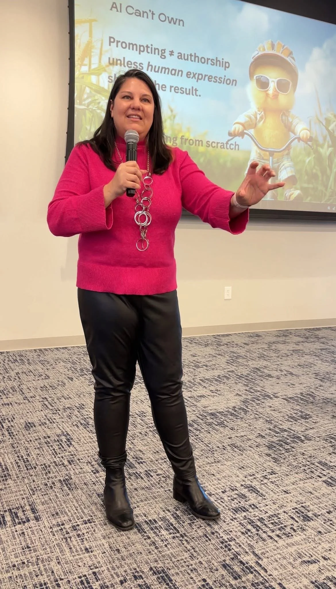 A woman in a pink sweater and black pants giving a presentation with a microphone, standing in front of a slide with a cartoon character of a pumpkin wearing sunglasses, against a background of cornfields.