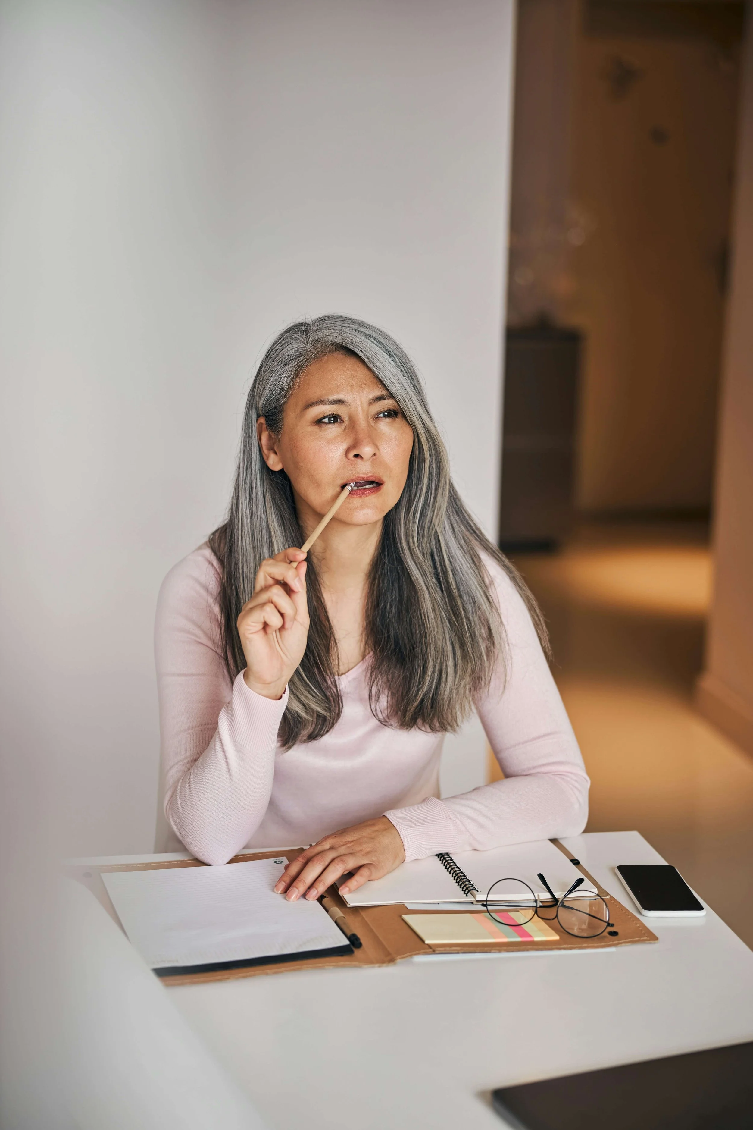 silver haired Asian woman with pencil in her mouth sitting at desk with notebooks