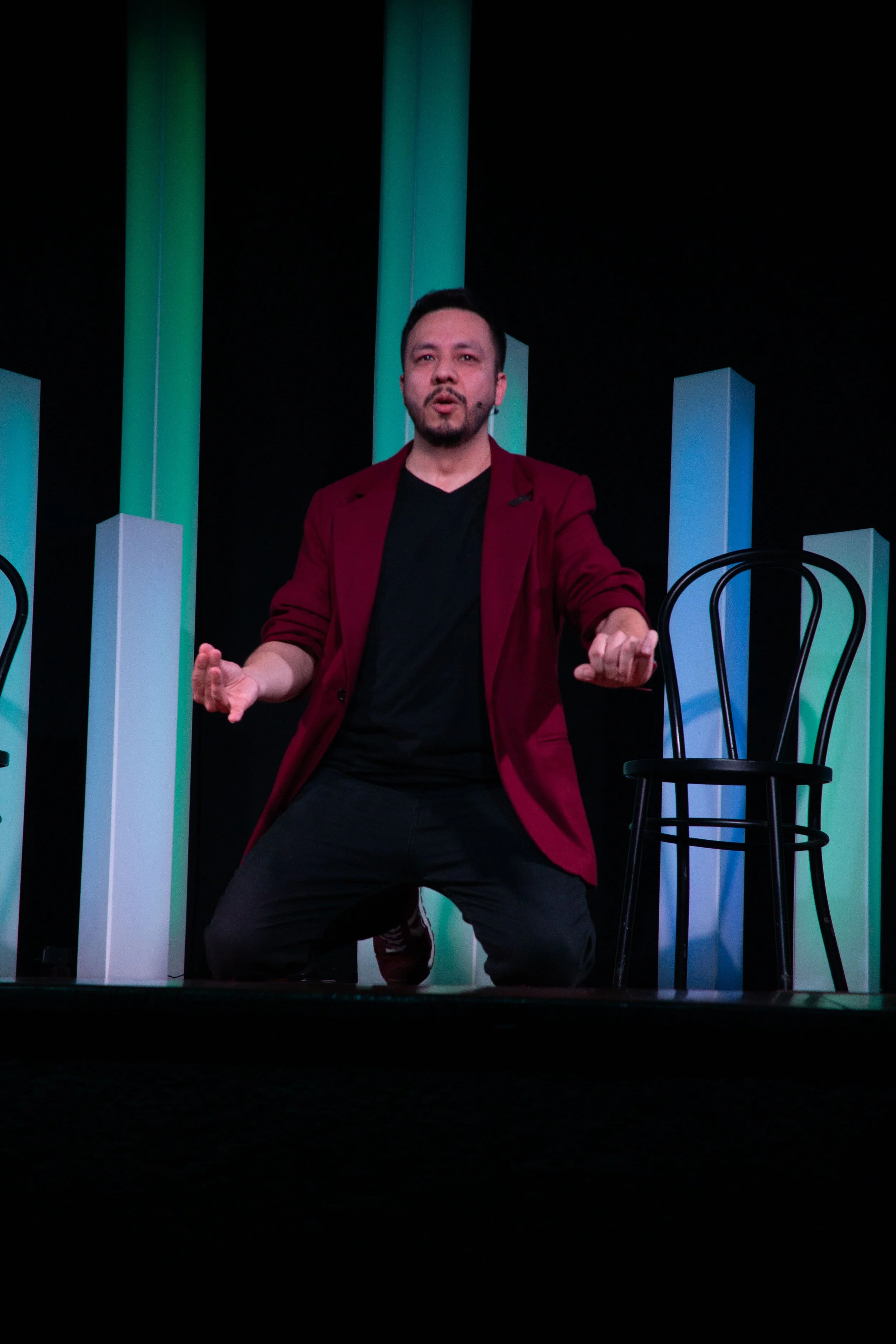 Juancho Carbono actuando en una obra de teatro, usando camiseta negra y saco rojo, en un escenario con estructuras blancas y verdes