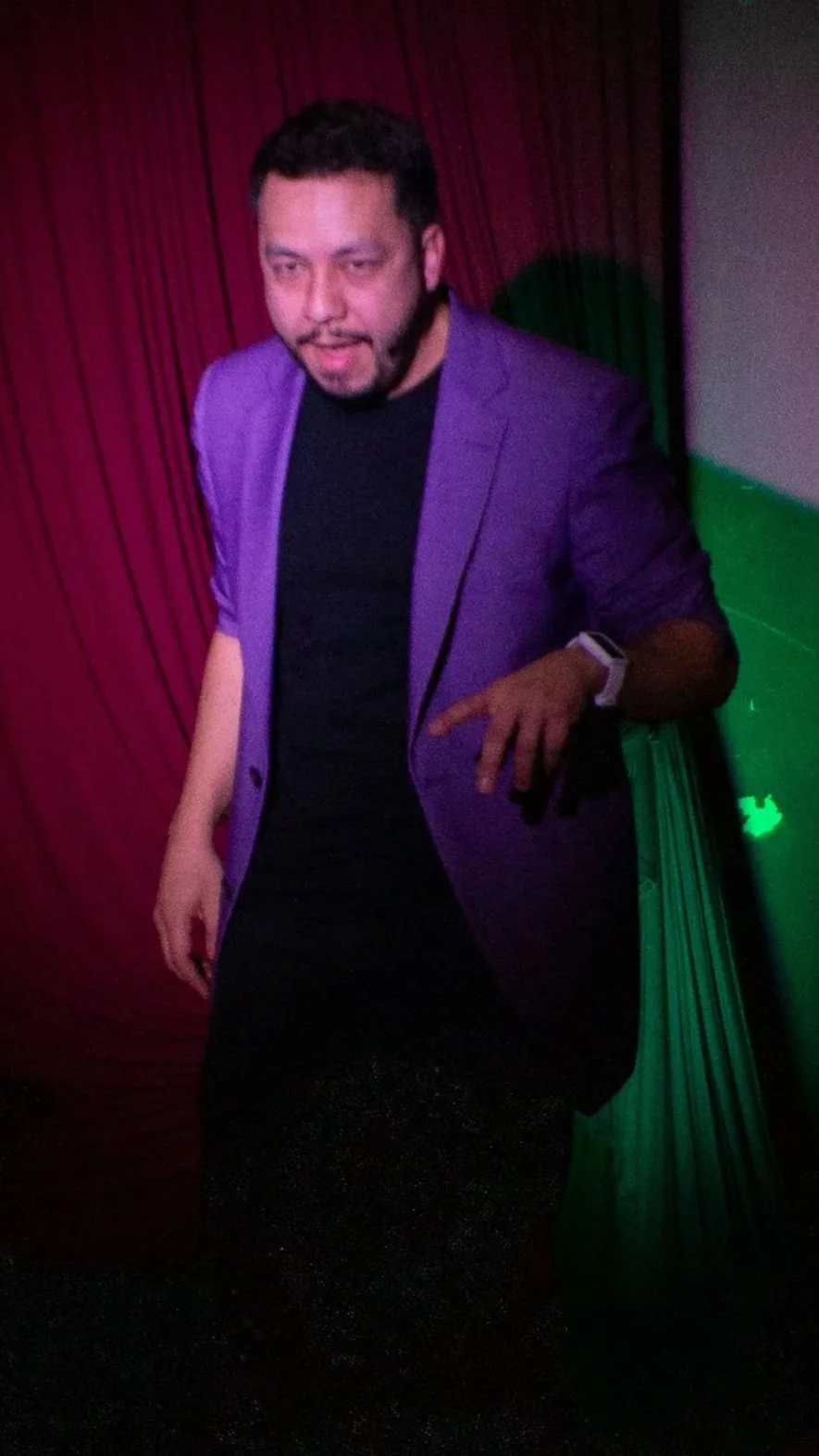 Juancho Carbono, vistiendo saco morado y camiseta negra en un lugar con cortinas rojas y paredes verdes, iluminado con luces de discoteca.