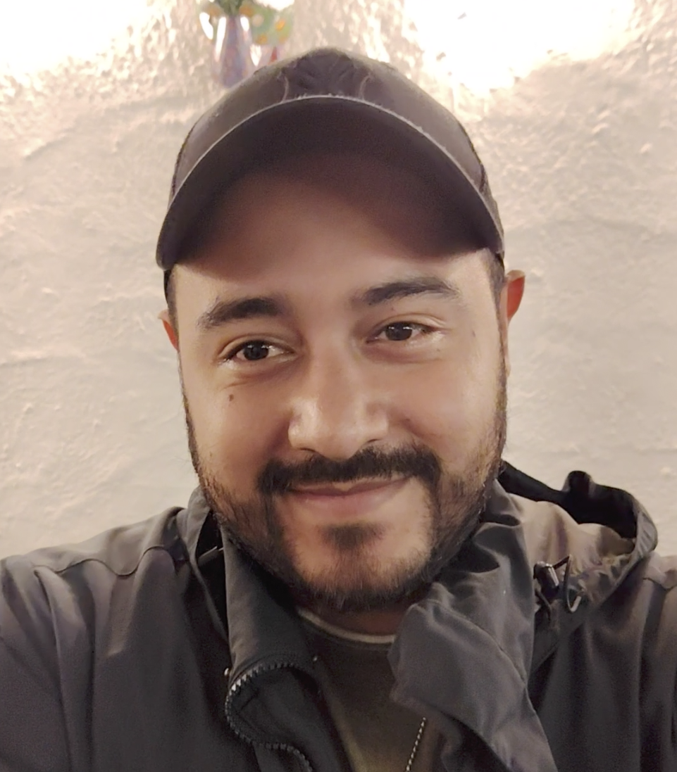 Primer plano de un hombre con barba, barba, usando una gorra negra y una chaqueta gris, sonriendo frente a una pared de color claro.