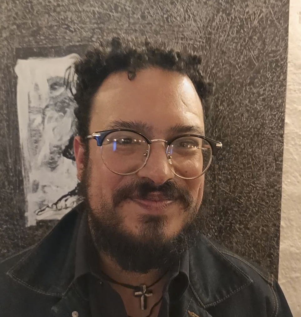 Un hombre con barba, lentes y collar con cruz, sonriendo y posando frente a una pared de color oscuro con una caricatura en blanco y negro estampada en ella.