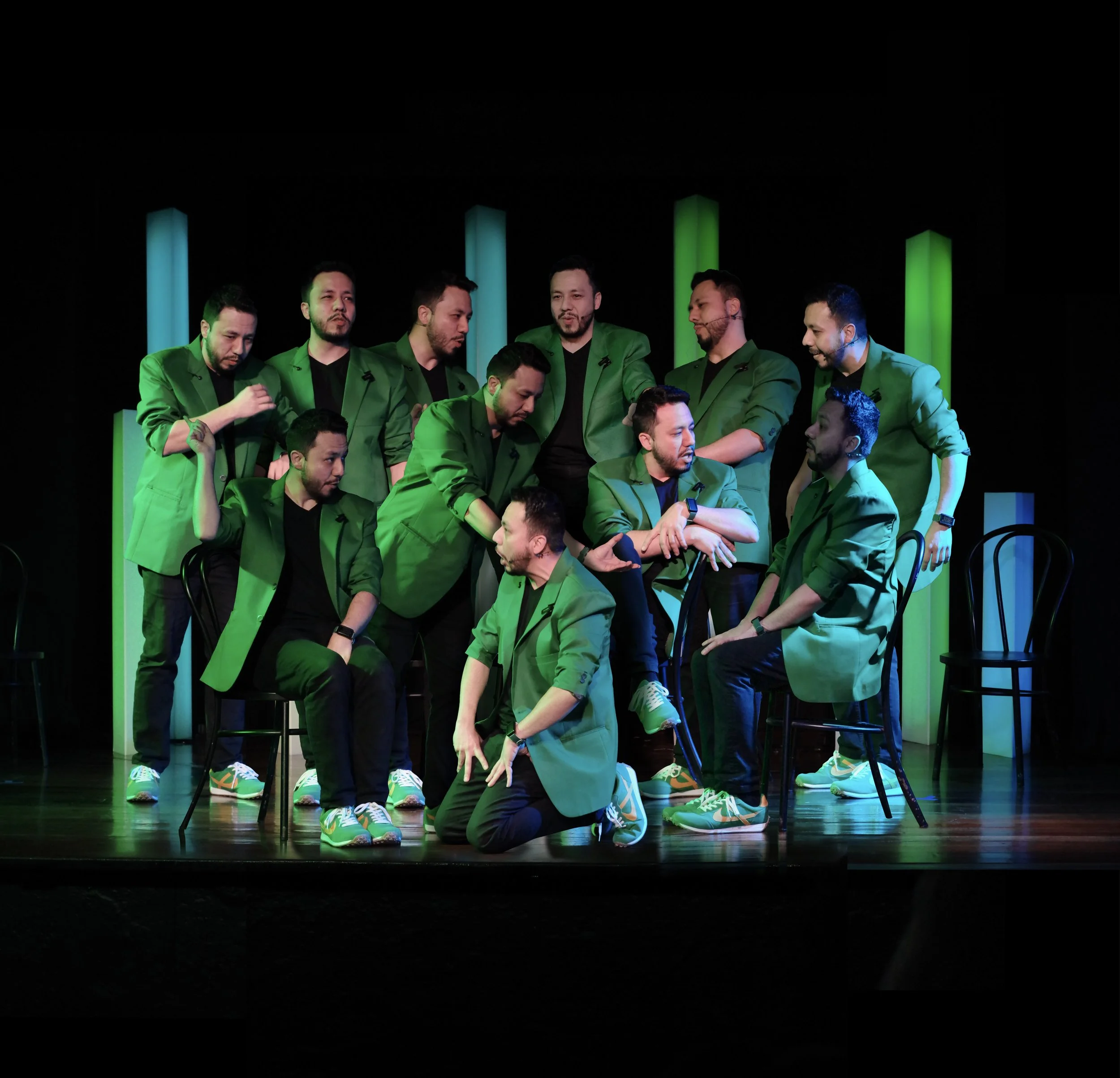 Múltiples copias de Juancho Carbono, todos en trajes verdes en un escenario, simulando una escena teatral o de comedia, con luces verdes y fondo negro.