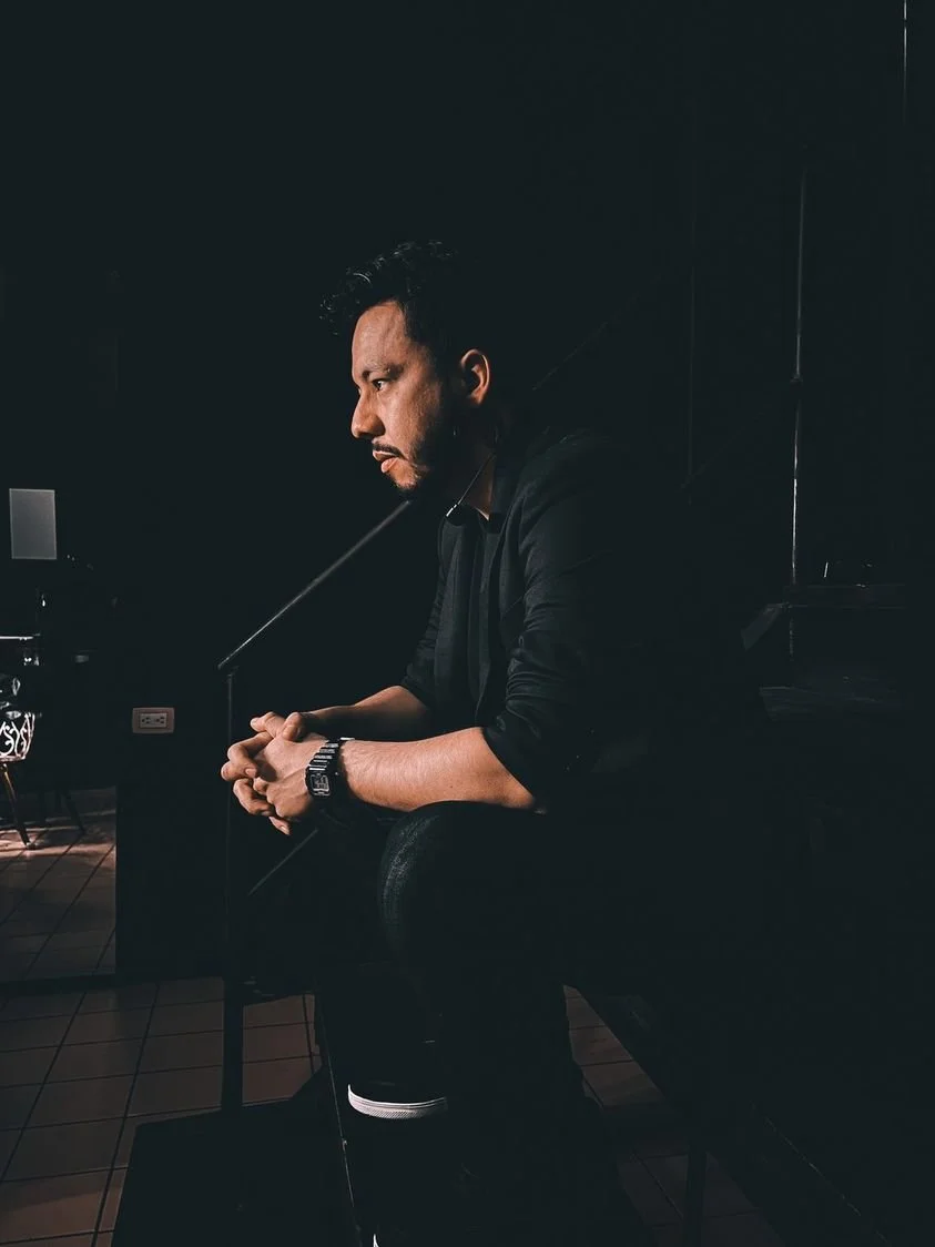 Juancho Carbono sentado en un ambiente oscuro, con expresión pensativa, con las manos juntas y viste ropa negra.