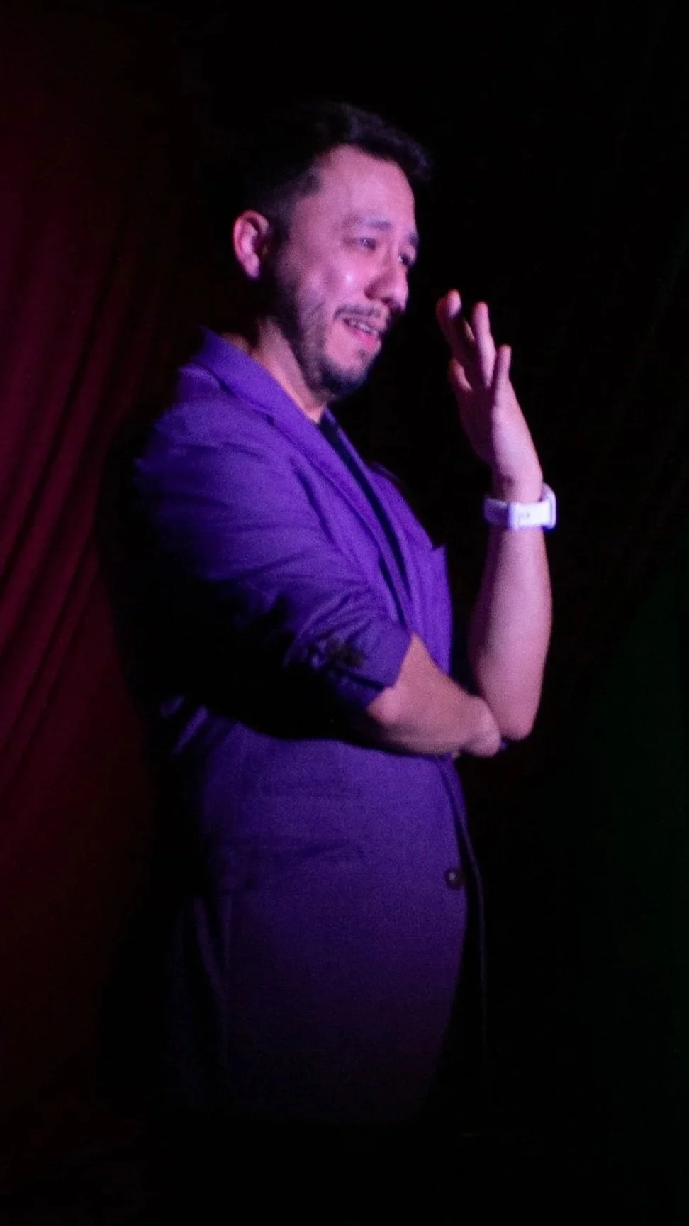 Juancho Carbono en escenario con expresión de tristeza o emoción, vistiendo saco morado, gesticulando con la mano y con fondo oscuro.