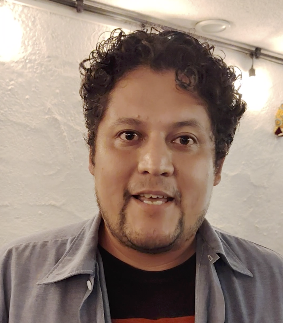 Un hombre con cabello rizado y barba, sonriendo, en un fondo interior con paredes blancas y una luz cálida.