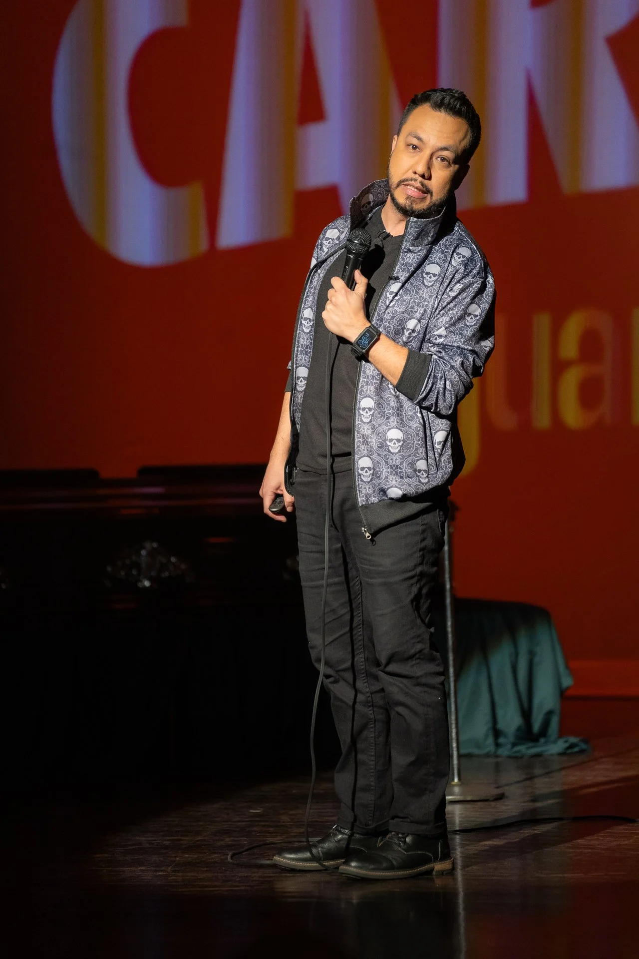 Juancho Carbono, comediante guatemalteco, en escenario de comedia, sosteniendo micrófono con fondo rojo y letras grandes.