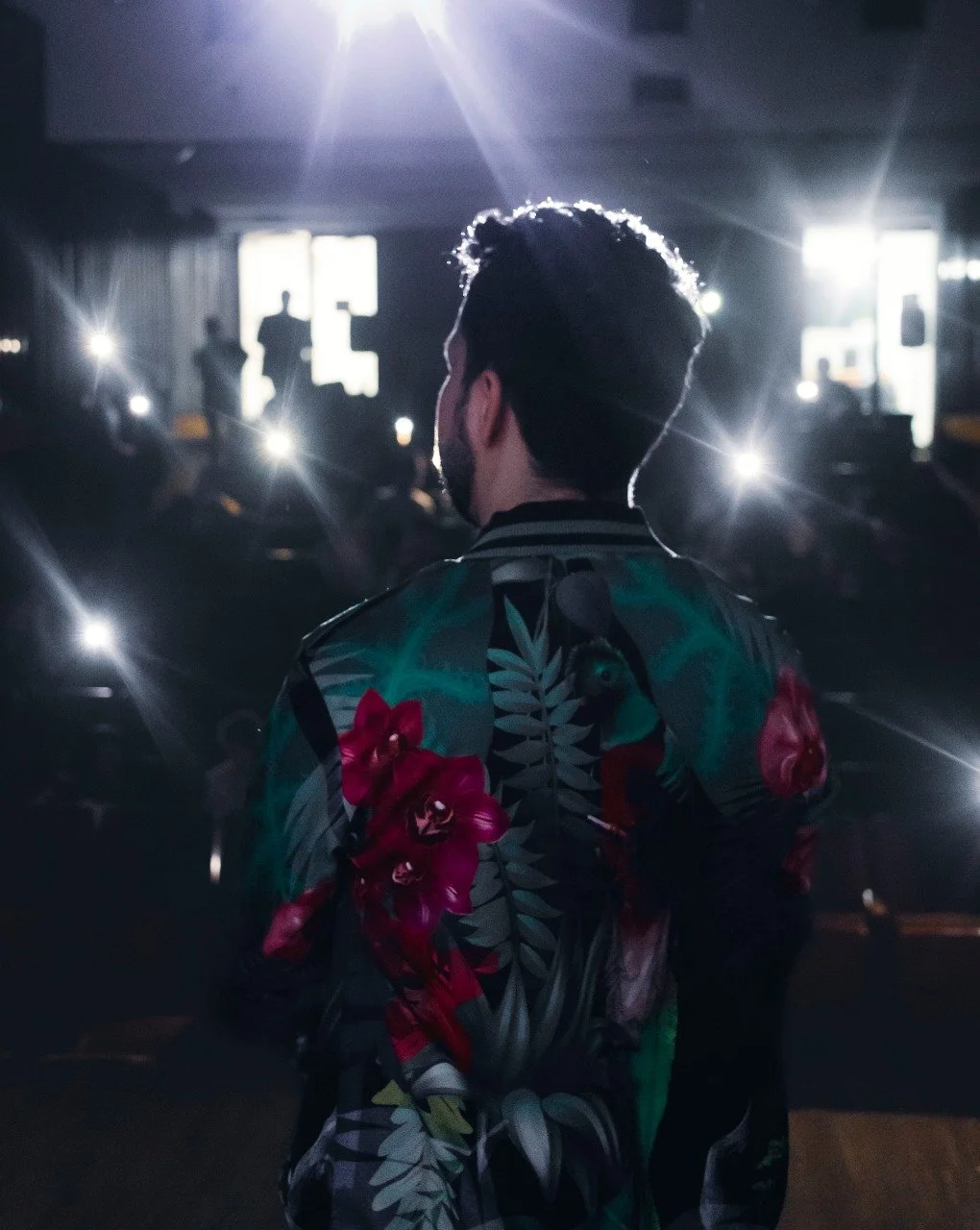 Juancho Carbono, comediante guatemalteco con chaqueta de colores oscuros y estampados de flores parado sobre el escenario en su show con luz brillante detrás y muchas personas con teléfonos en la mano