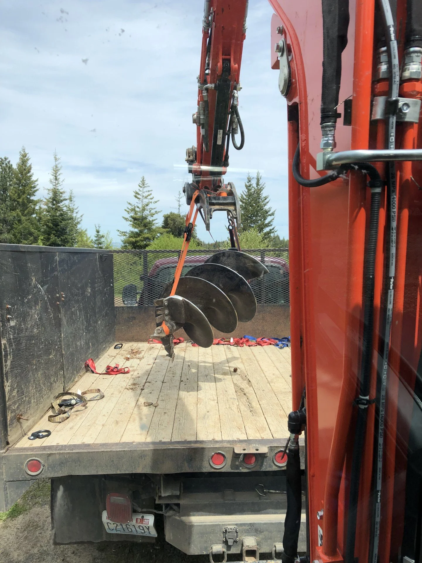 A truck with a large orange excavator arm, holding an auger drill bit used for digging holes, parked outdoors on a partly cloudy day with trees in the background.
