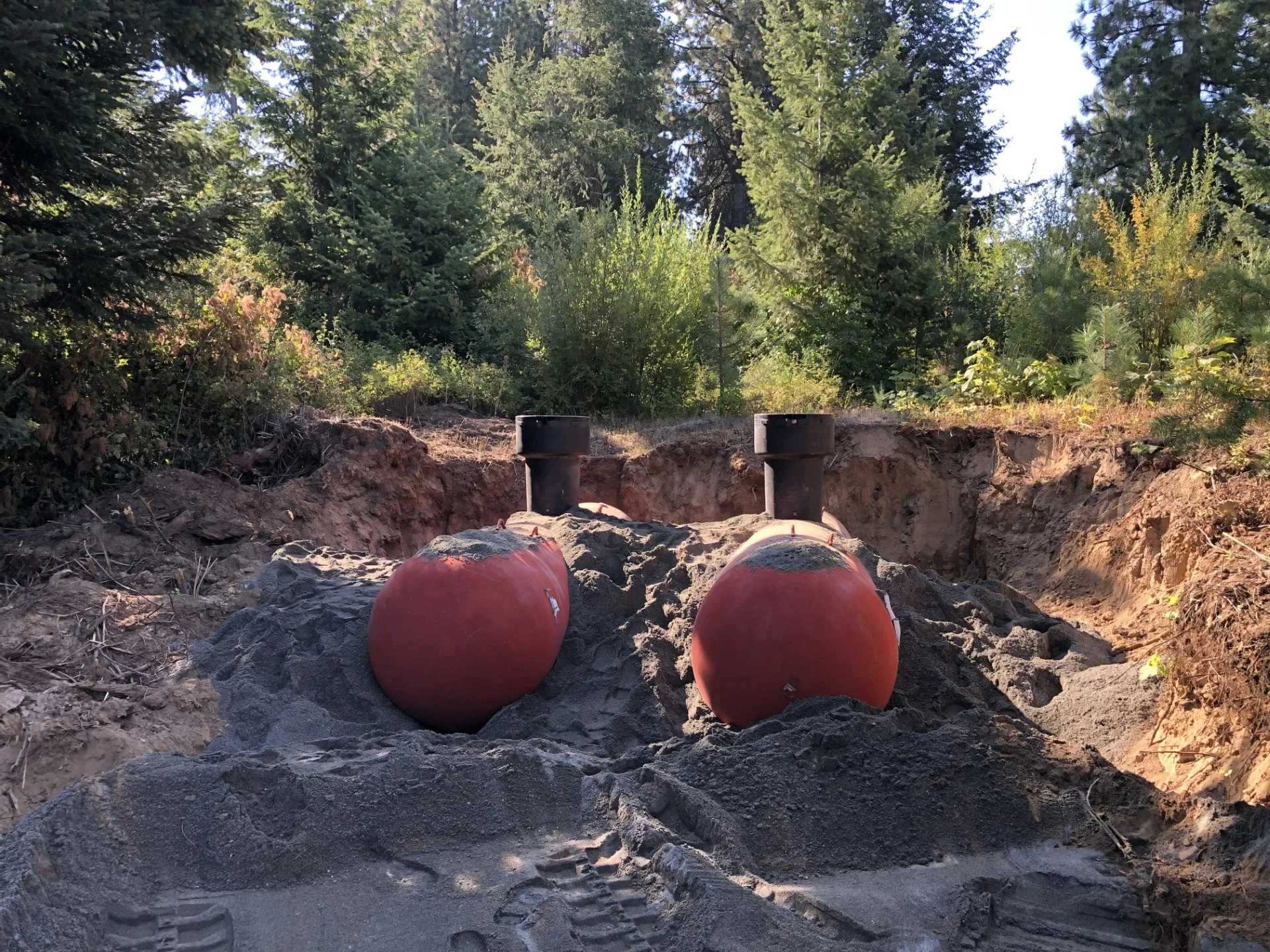 Two buried devices with black tops in a dirt excavation site surrounded by trees and bushes.