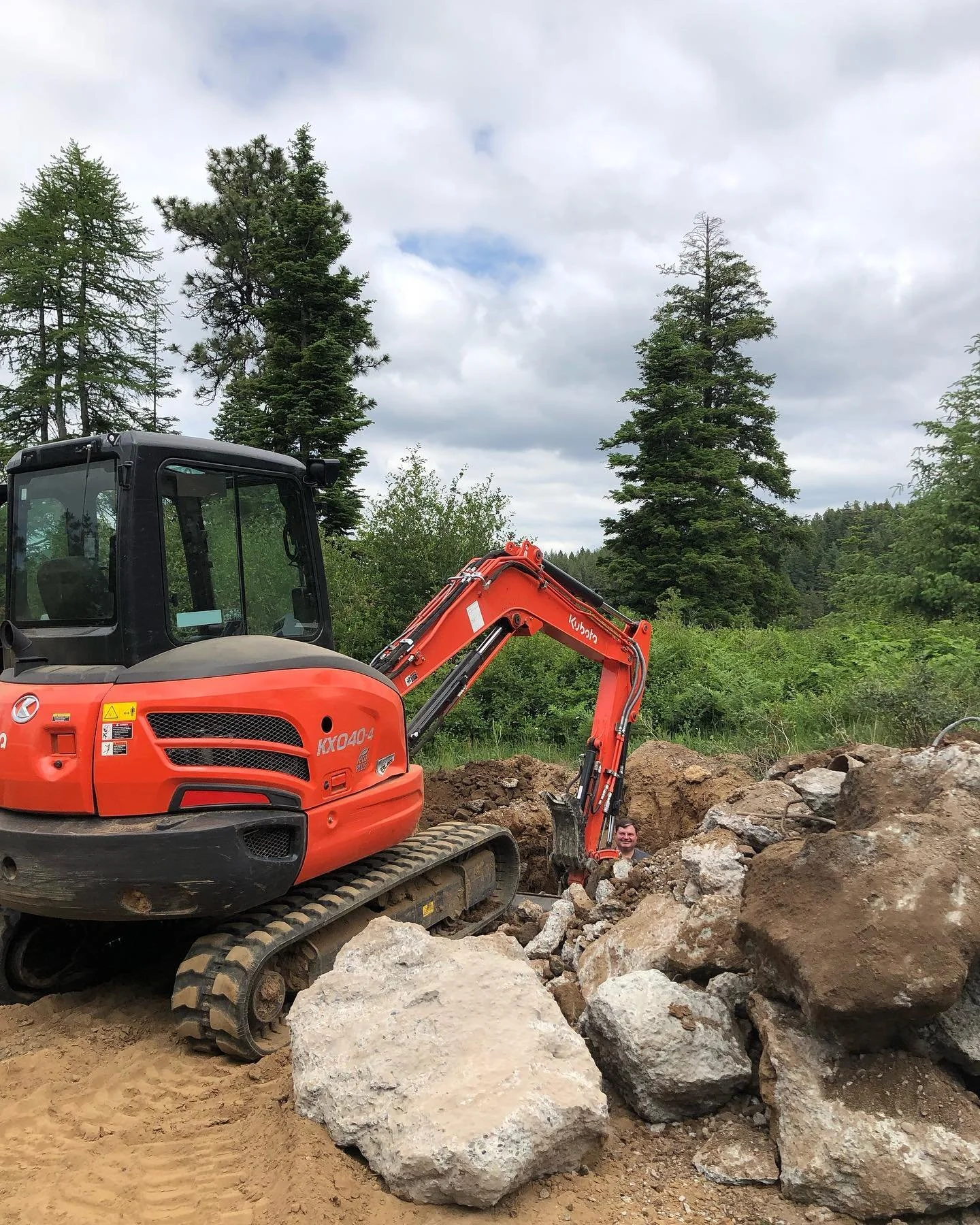 A small orange excavator working on a dirt construction site surrounded by rocks and green trees with a cloudy sky overhead.