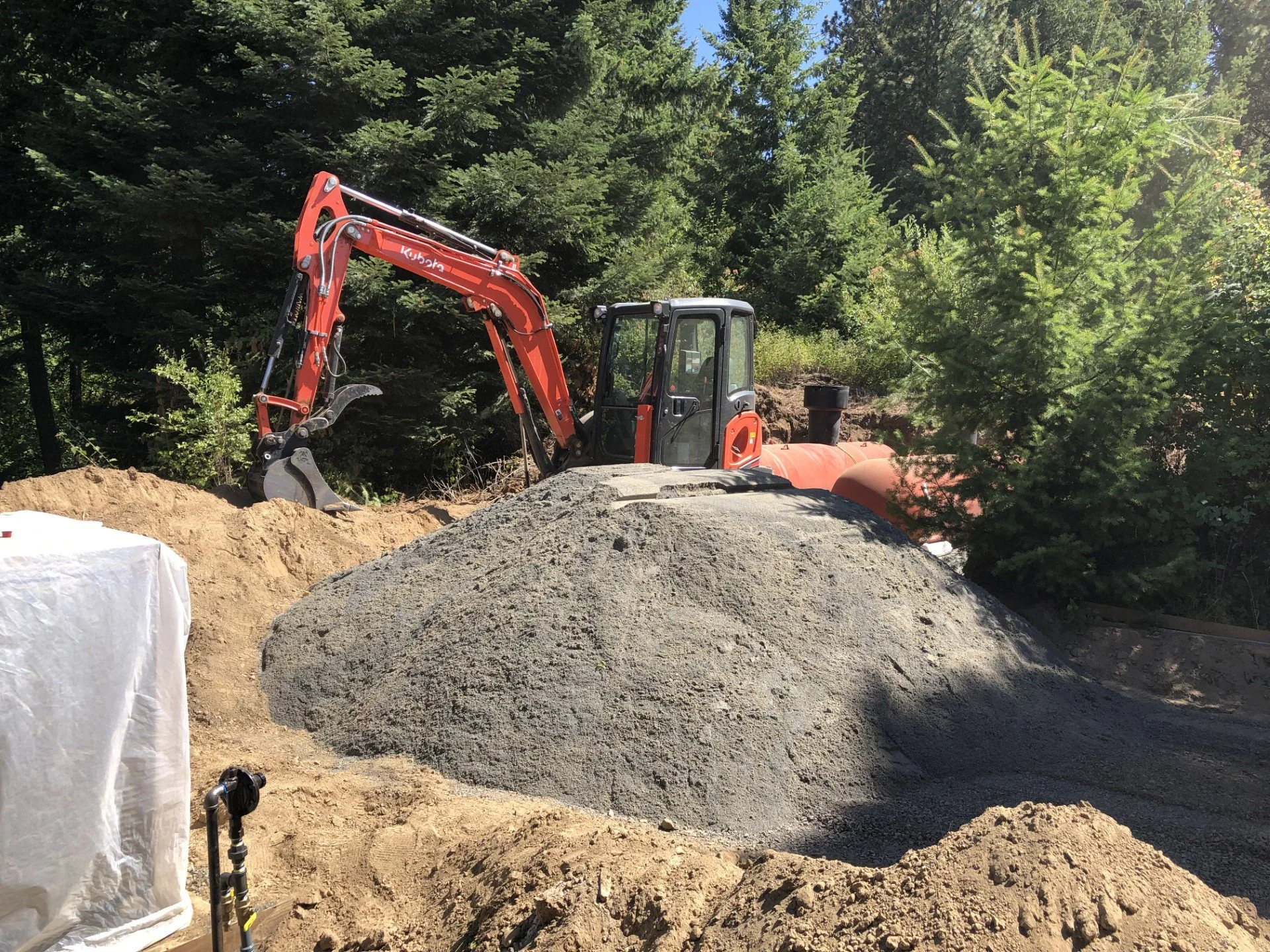 A red compact excavator working on a construction site with a large pile of gravel or dirt, surrounded by trees and greenery.