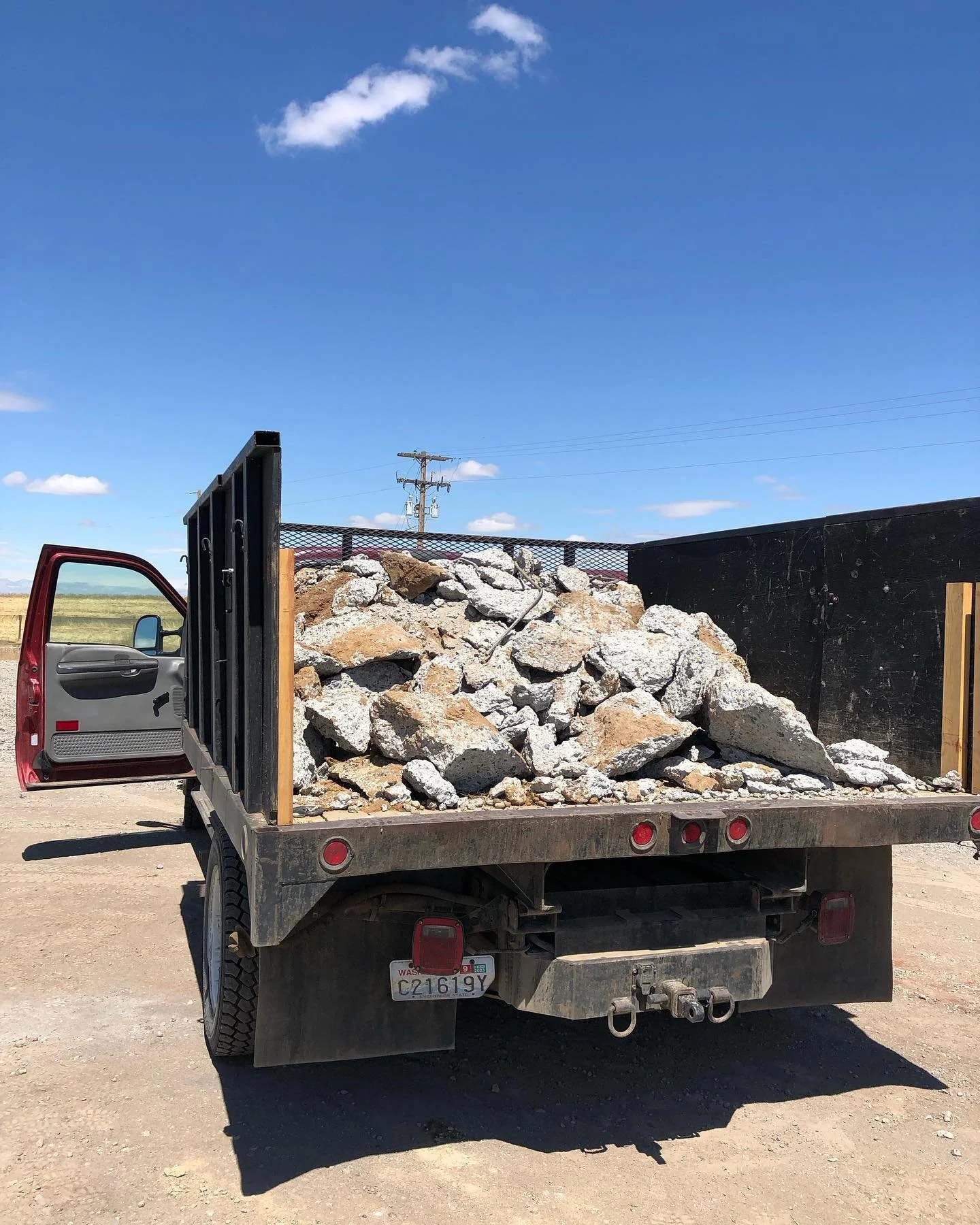 A flatbed truck loaded with rocks and debris parked on a dirt surface with open land and utility poles in the background under a blue sky with a few clouds.