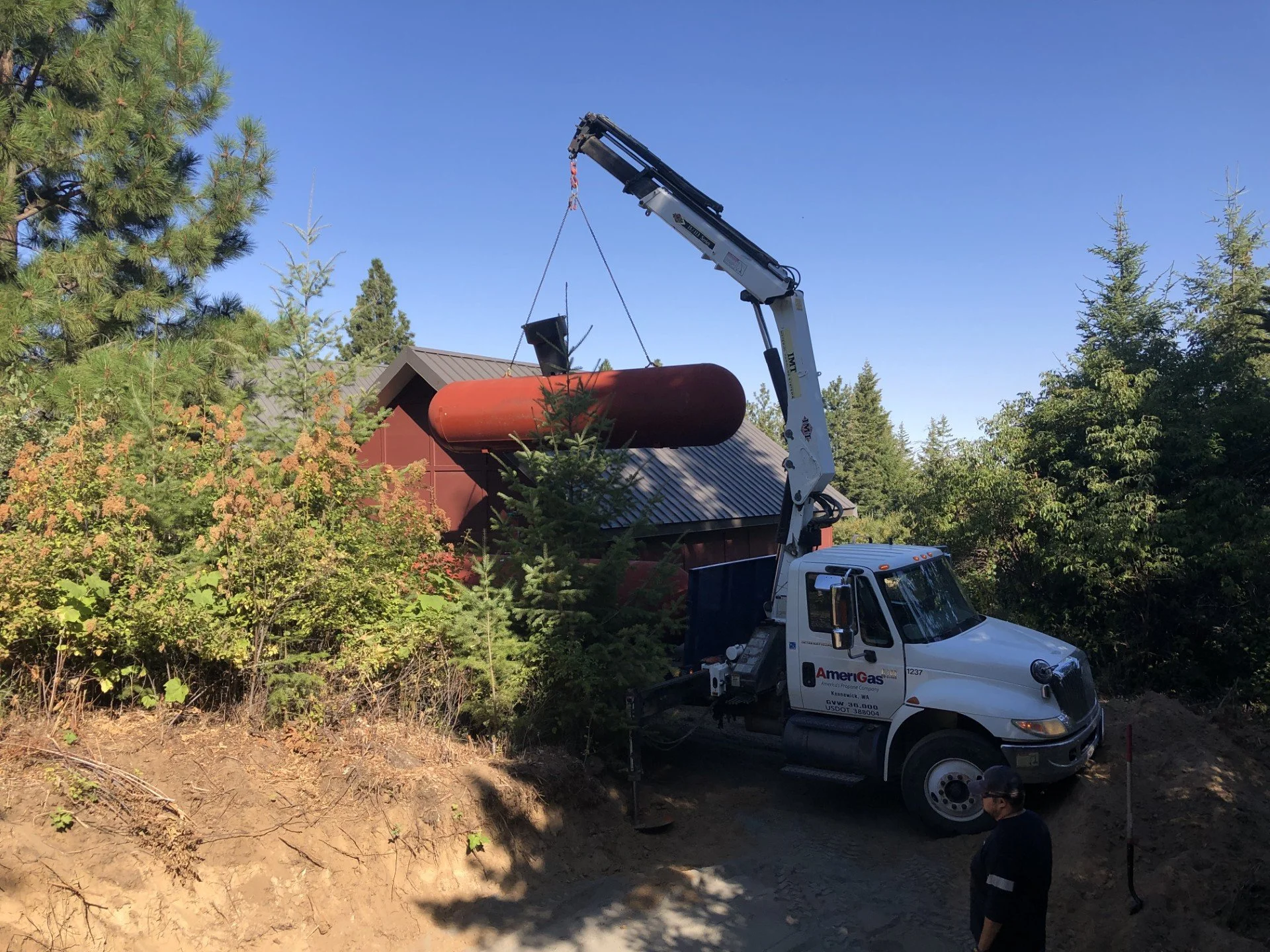 A utility truck with a mounted crane lifting a large tank into a building surrounded by trees under a clear blue sky.