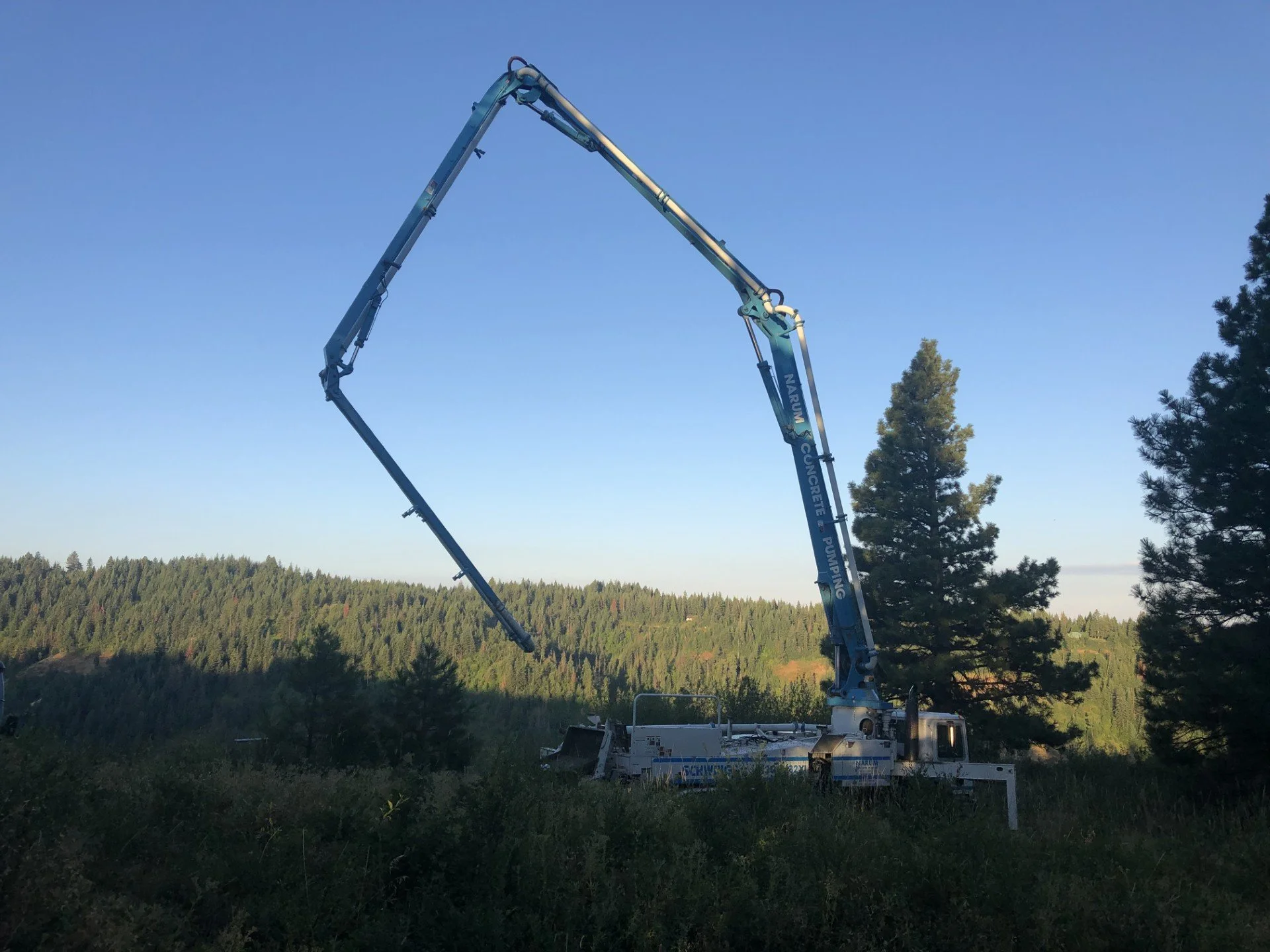 A concrete pump truck with a long, extendable boom arm is situated in a grassy area with trees and a forested hillside in the background.