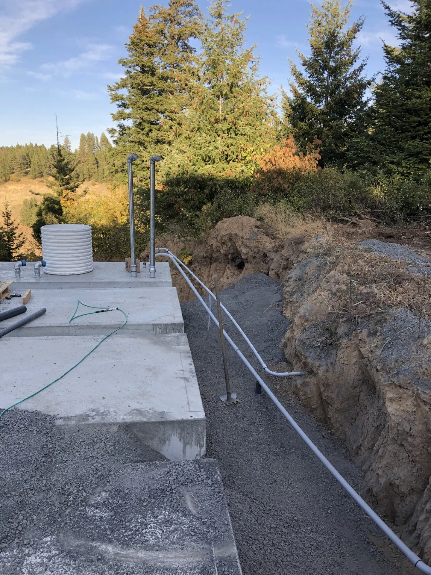 Construction site with concrete foundation, plumbing pipes, and electrical conduits, surrounded by dirt and gravel, with trees and a clear sky in the background.